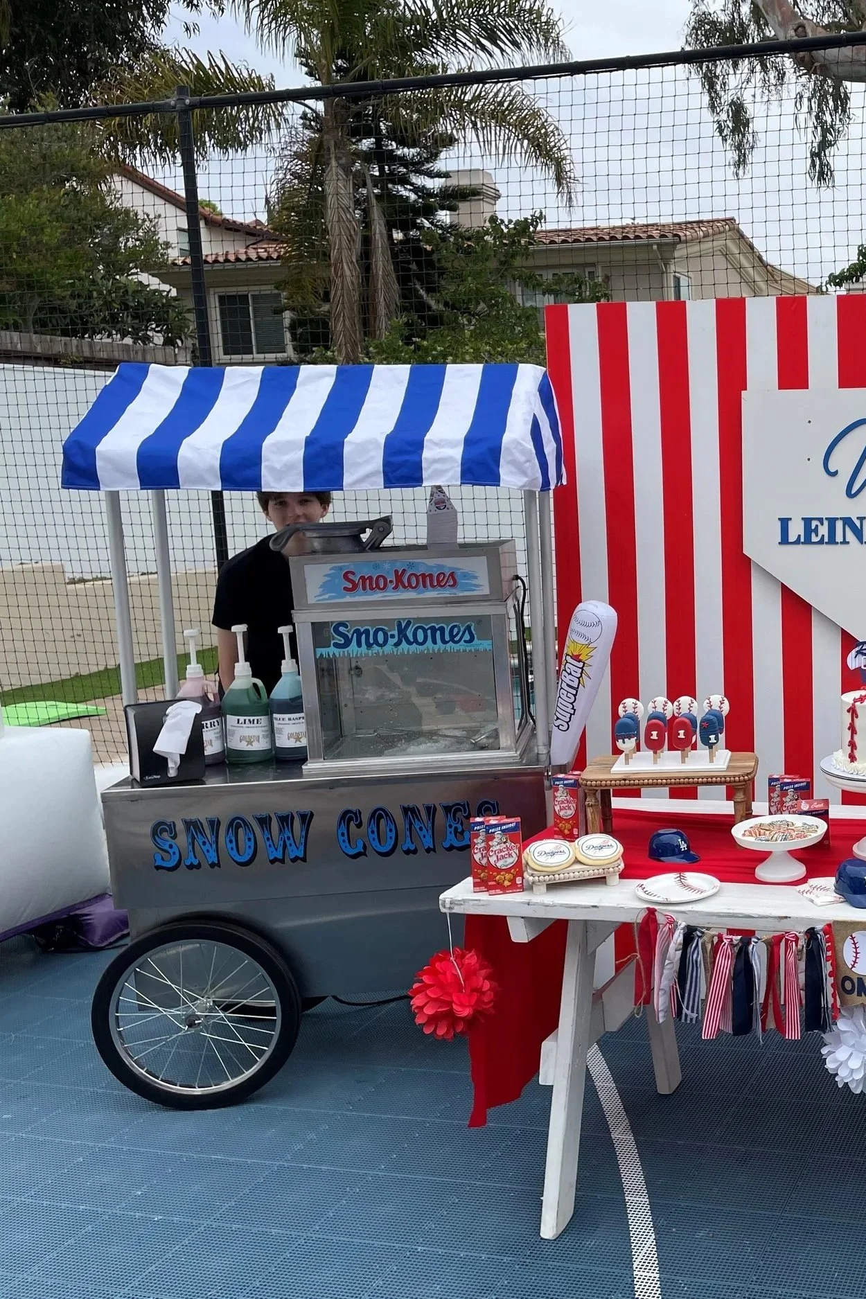 A boy selling snow cones at a stall decorated with a blue and white striped canopy at an outdoor event, with a red, white, and blue striped backdrop and baseball-themed decorations.