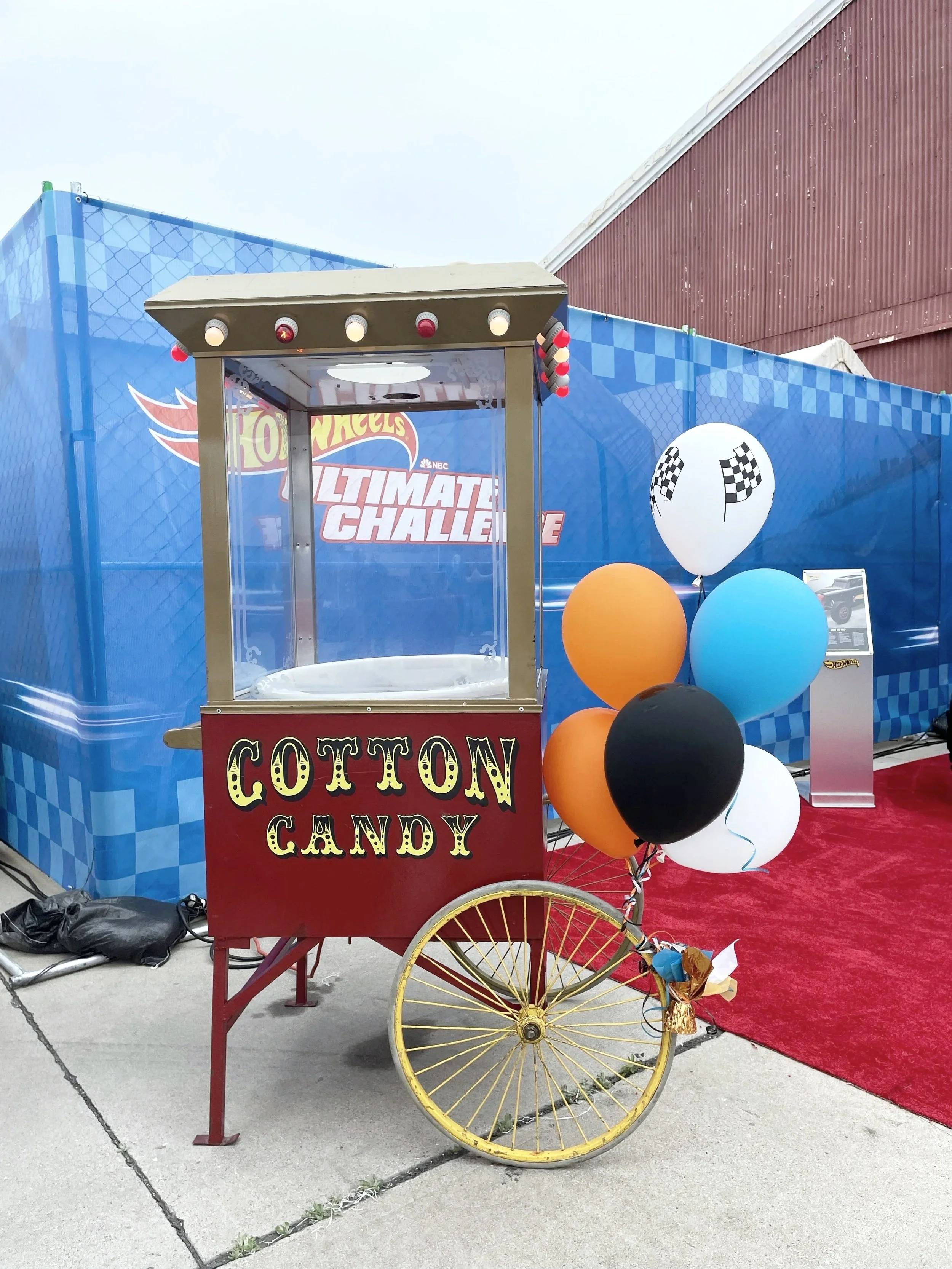 A vintage cotton candy cart with yellow wheels, red frame, and an attached bunch of balloons in orange, black, white, and blue. The cart has a sign reading 'COTTON CANDY' in retro style lettering. Behind the cart is a blue checkered branded backdrop with balloons nearby.