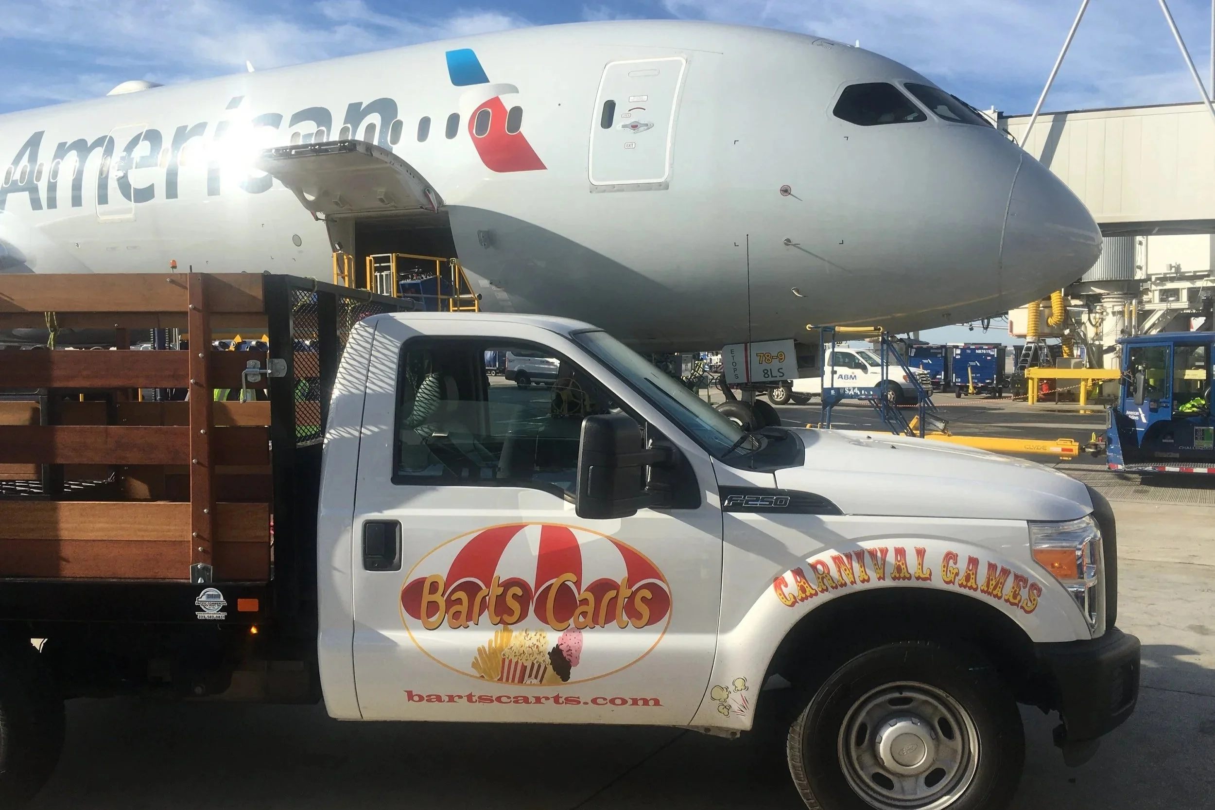 A white pickup truck with a tray carrying a wooden crate with the logo 'Barts Cars' in front of an airplane at an airport gate.