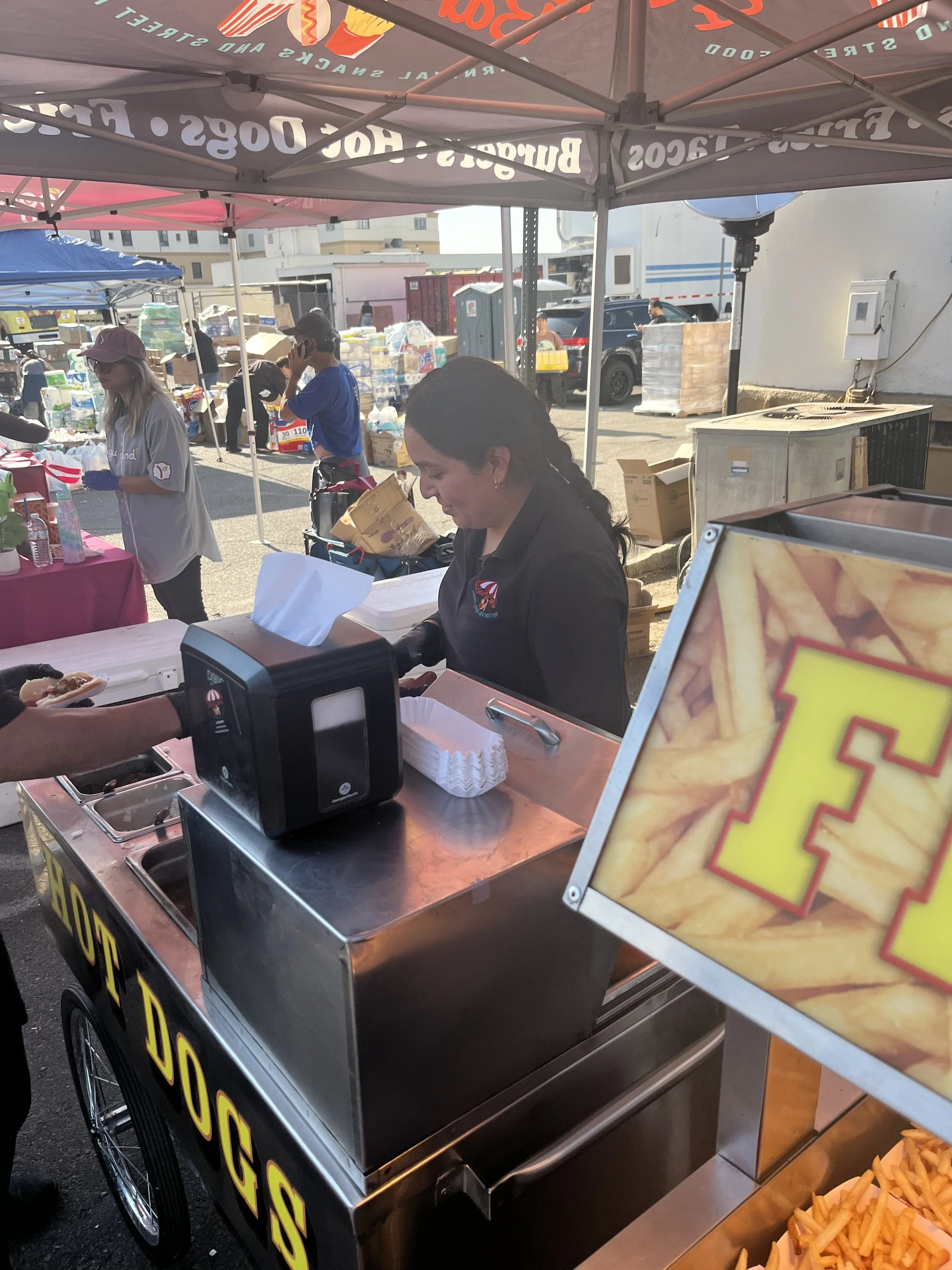 A mobile food stand selling cheese fries, with a sign displaying a picture of cheese fries and a large yellow and red 'E' on it. There are people behind the stand and in the background, some with shopping carts, under tents at an outdoor market.