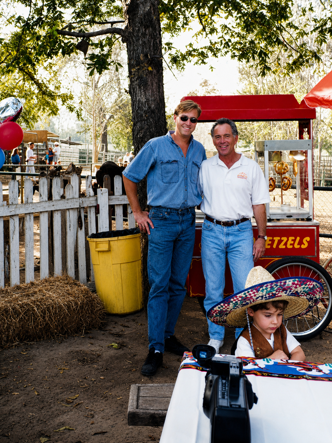 Two men standing outdoors by a red popcorn and pretzel cart labeled 'Pretzels,' one wearing sunglasses and a denim shirt, the other in a white polo shirt. A young boy with a sombrero is sitting at a table in the foreground, with a camera pointed at the scene. There are trees, a white picket fence, and a few people in the background.