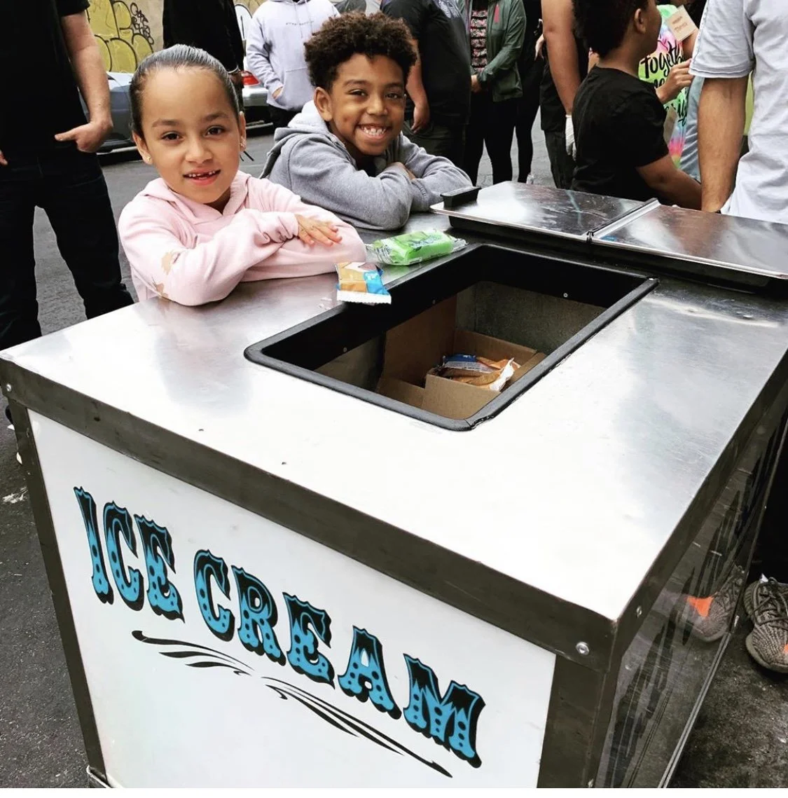 Two children smiling and standing at an ice cream stand, which is open with the lid lifted, showing some items inside. There are several people in the background.