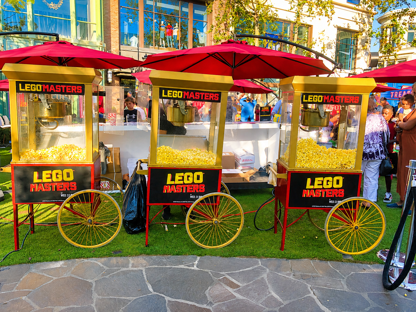A street vendor cart selling LEGO-shaped popcorn in three separate compartments, with a crowd of people around and a modern building with large glass windows in the background.