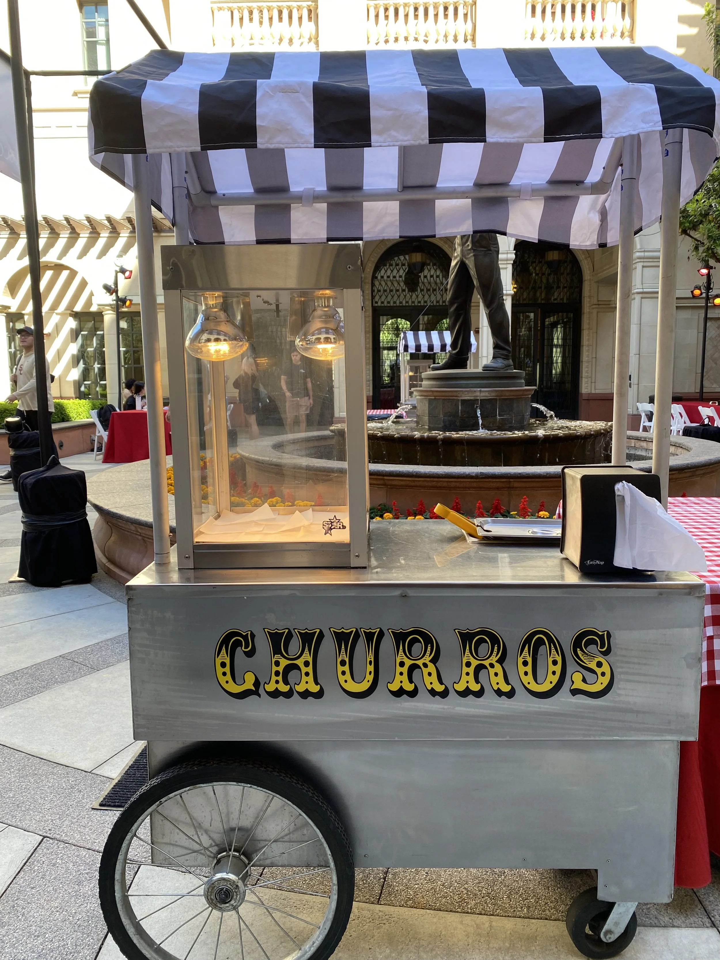 A food cart selling churros with a striped black and white canopy and a sign that reads "Churros." The cart has two wheels and a metal body, with a display area and a serving window. In the background, there are people, outdoor seating, and a fountain.