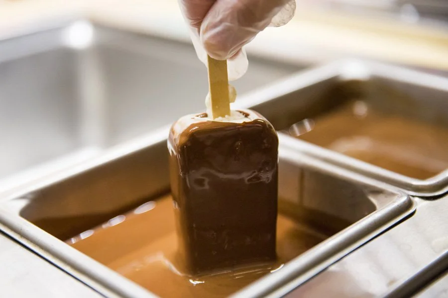Close-up of a hand wearing a glove holding a wooden stick, dipping a chocolate-coated ice cream bar into a container of melted chocolate.