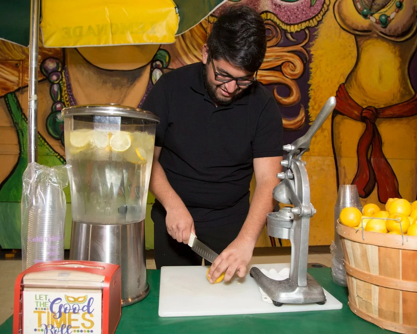 A man slicing a lemon at a stand with a basket of lemons and a lemonade dispenser.