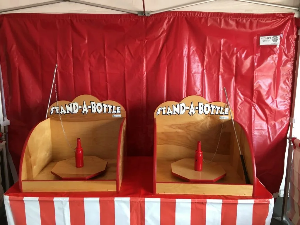 Two wooden stand-a-bottle game booths with red and white striped fabric at the front, each featuring a red bottle on a small table and a sign that reads 'STAND-A-BOTTLE' at the top, set against a red backdrop.