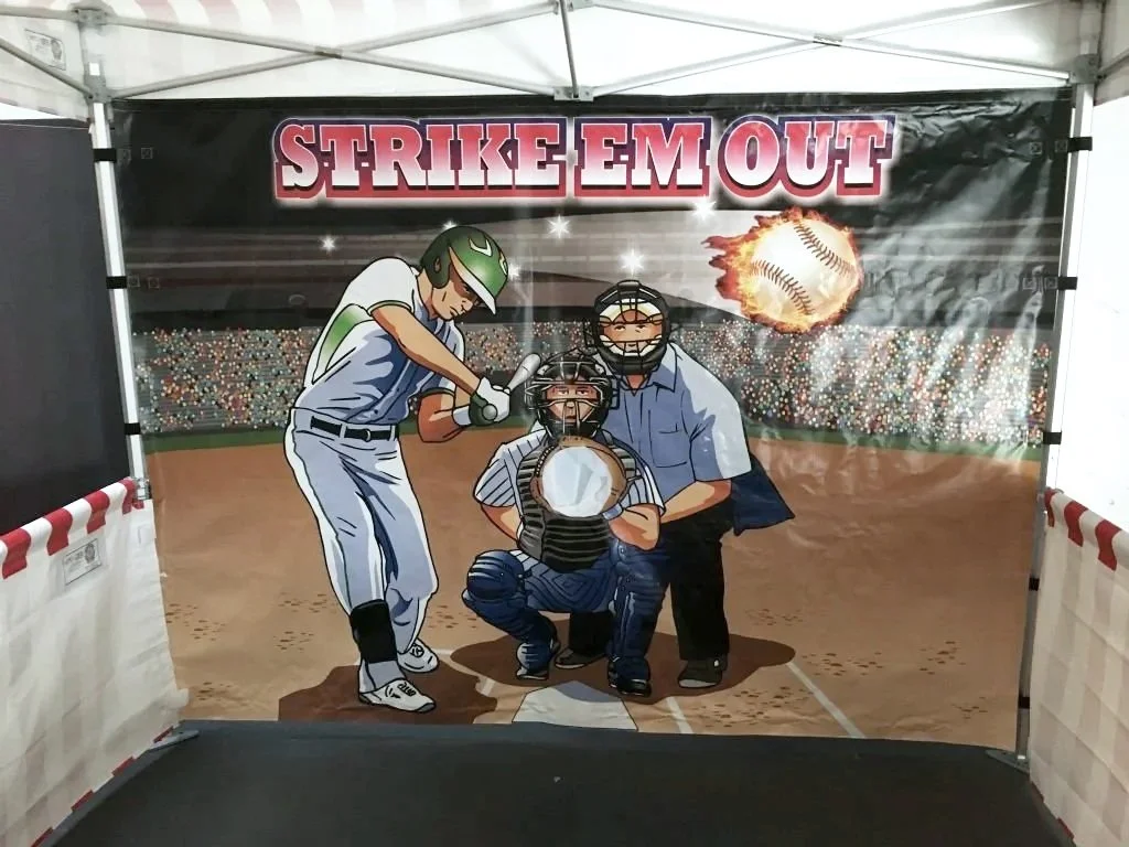A baseball-themed game booth with a cartoon depiction of a batter swinging, a catcher behind home plate, and an umpire to the side. The background shows a stadium with a large fiery baseball. Above, the sign reads 'STRIKE EM OUT' in bold red and black letters.