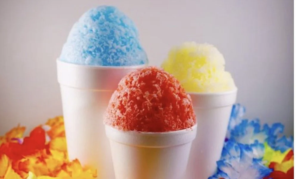 Three cups of shaved ice with different colored flavors: blue, yellow, and red, surrounded by colorful shredded paper.