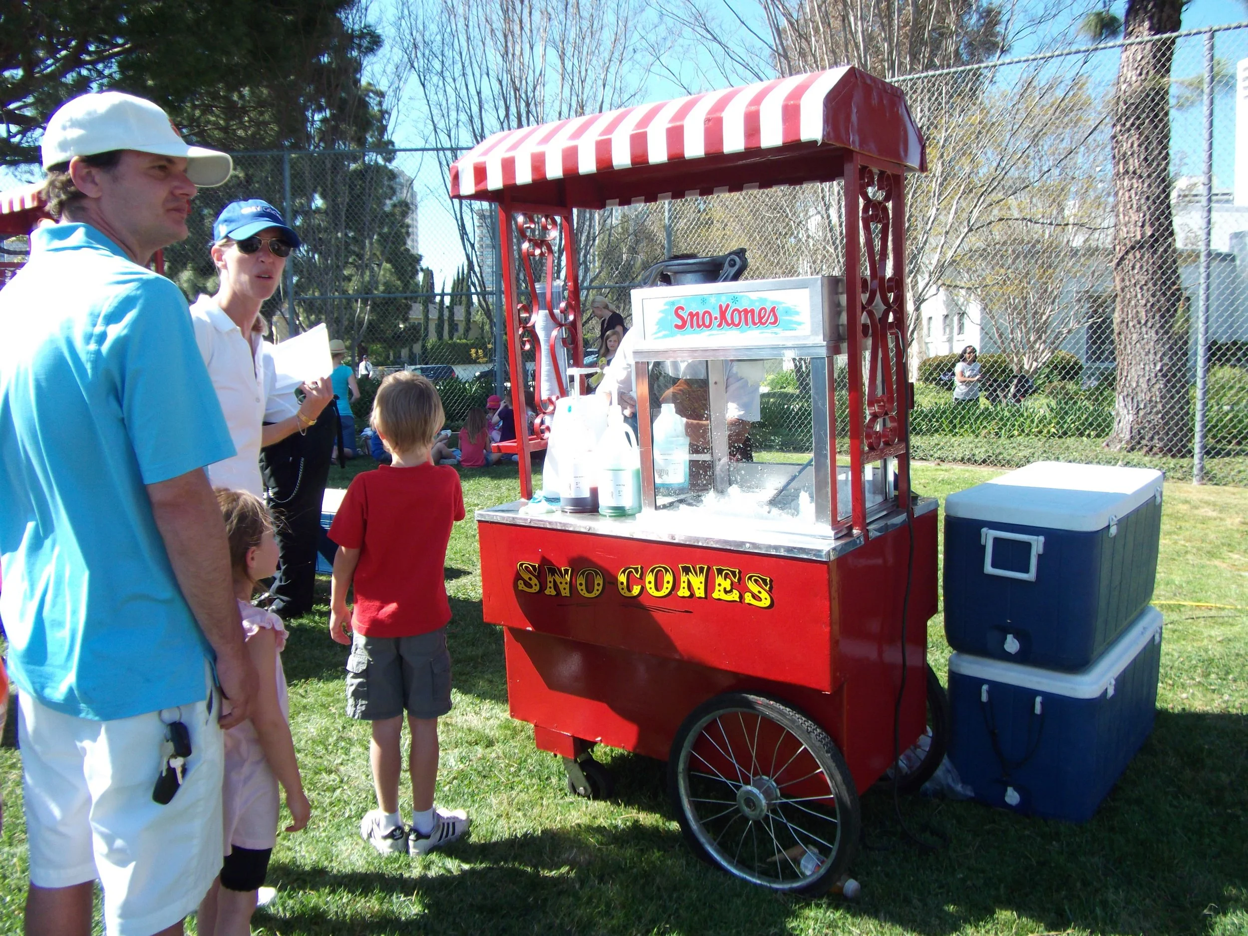 A group of people gathered outdoors around a red snow cone cart labeled 'Sno-Cones,' with a red and white striped canopy, on a sunny day in a park or recreational area surrounded by trees and a chain-link fence.