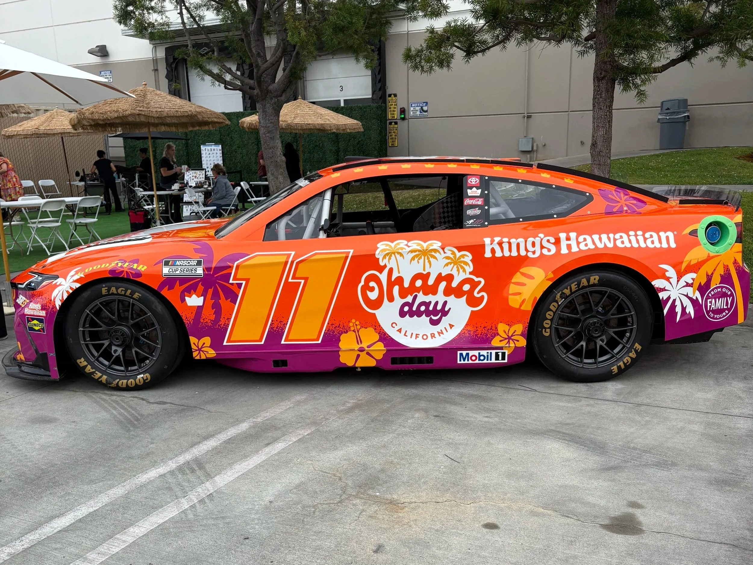 Colorful race car with Hawaiian-themed decals, parked in a lot near a grassy area with trees, umbrellas, and people at outdoor tables.
