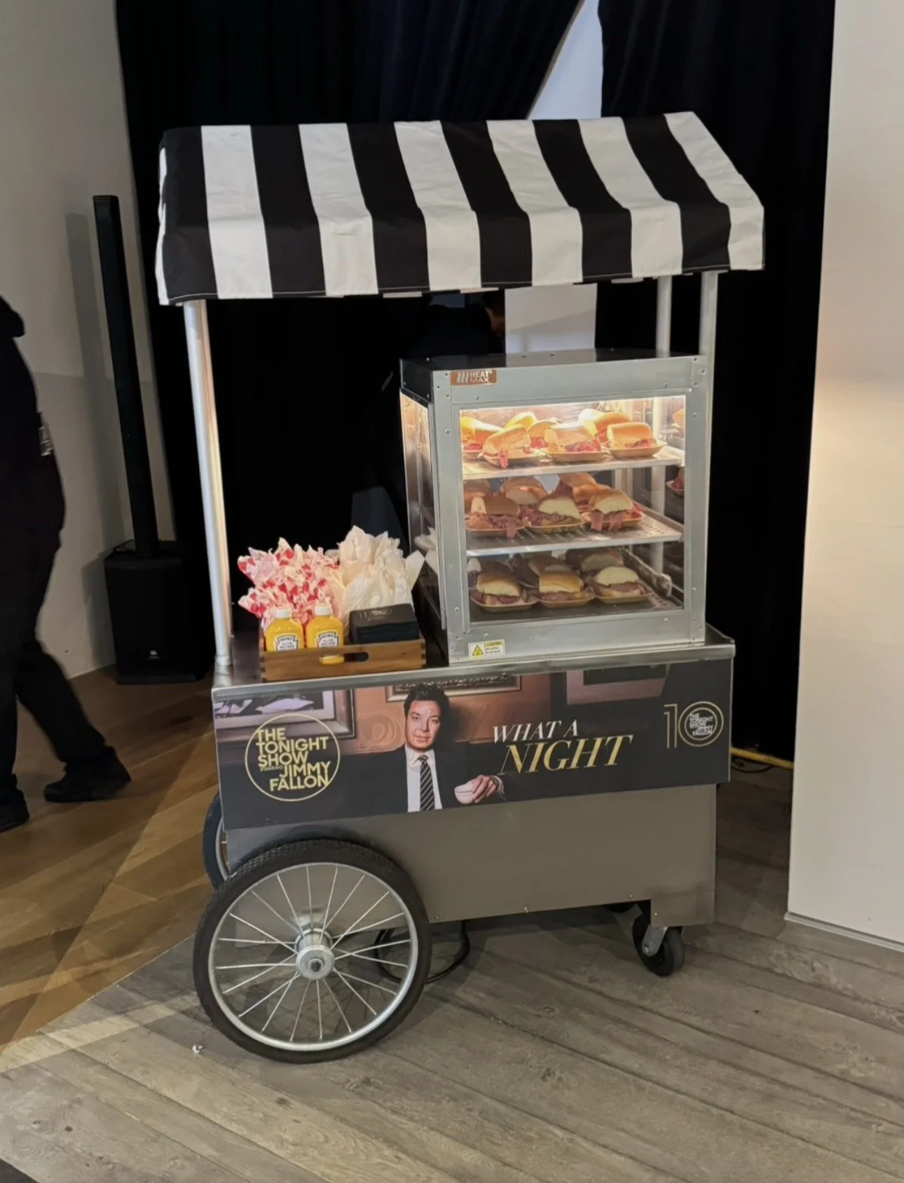 Mobile food cart selling sandwiches and hot dogs, with a black and white striped canopy, featuring promotional poster for 'The Tonight Show with Jimmy Fallon'.