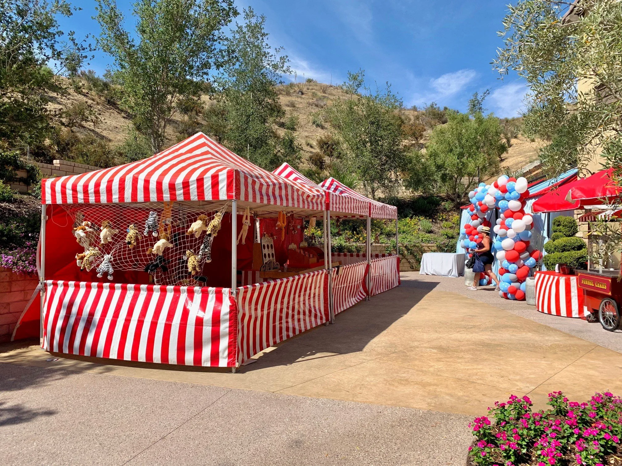Red and white striped tents at an outdoor fair with stuffed animal prizes, colorful balloons on the side, and a woman walking nearby against a hillside with trees and a clear blue sky.