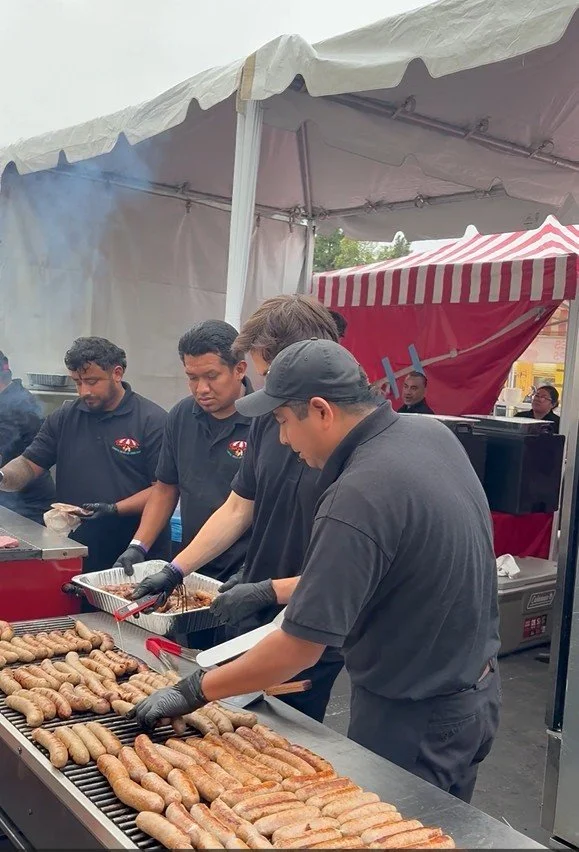 Four men cooking and grilling sausages at an outdoor event under a white tent with a red and white striped canopy in the background.