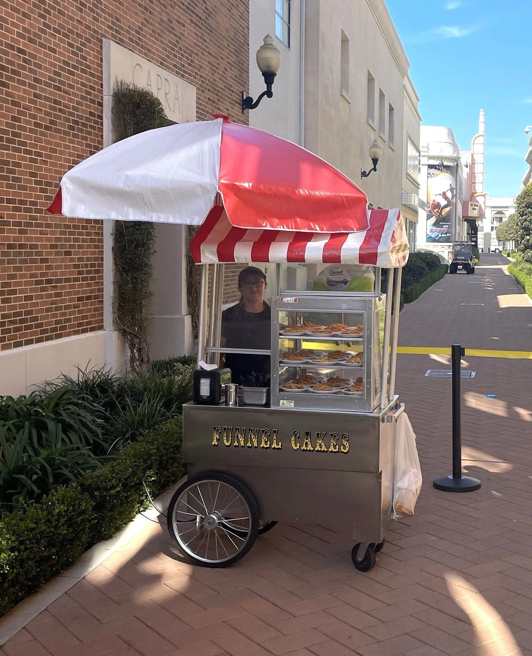 A food cart with a red and white striped umbrella parked next to a brick building and a sidewalk with hedges. The cart has a sign that reads 'FUNNEL CAKES' and displays plates of funnel cakes. A person is standing behind the cart. In the background, there are buildings, streetlights, and a billboard.