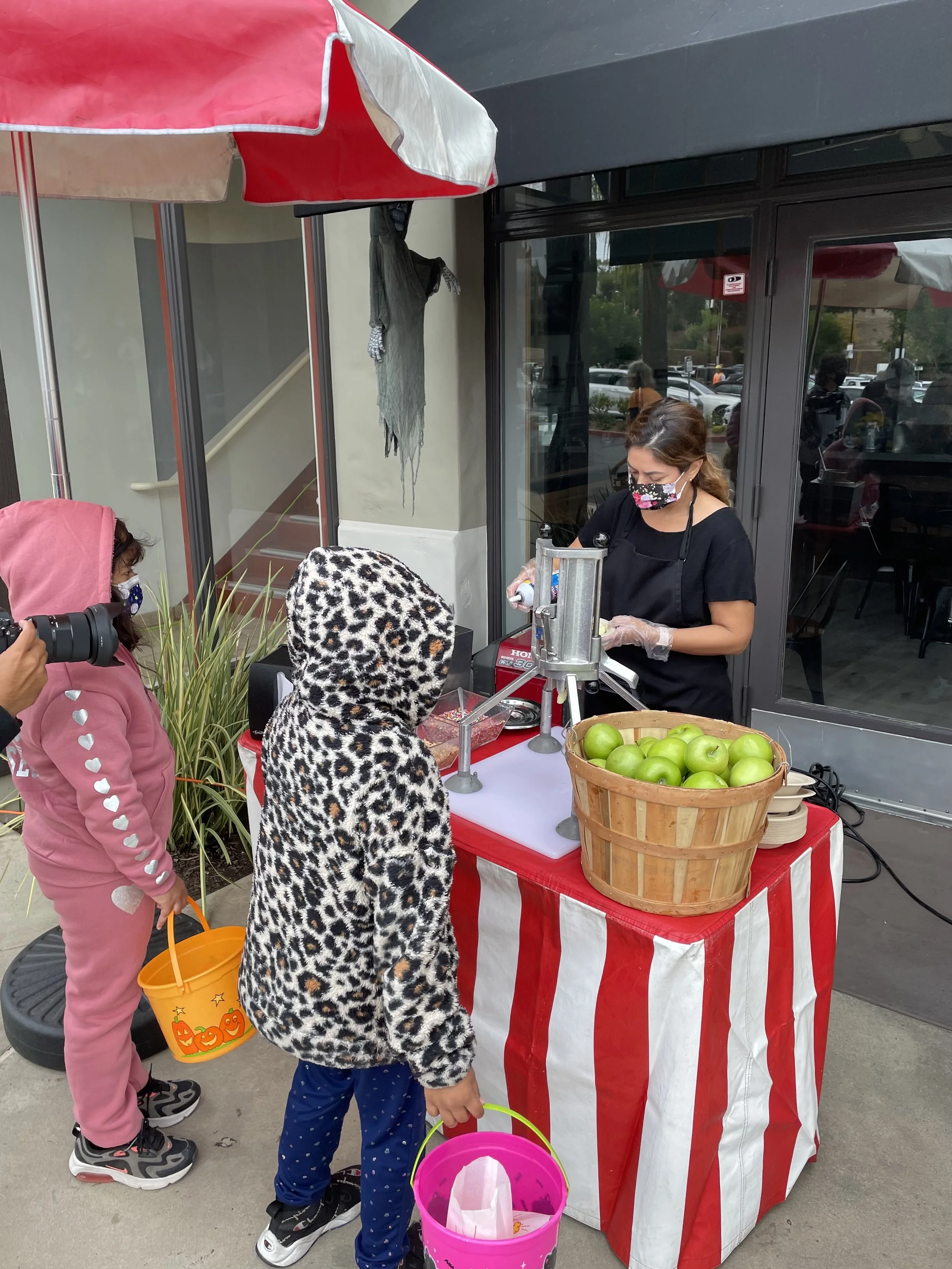 Children Trick-or-Treating at a Halloween event, collecting candy from a vendor's stand with a red and white striped table, a basket of green apples, and a woman handing out treats, all wearing Halloween costumes and masks.