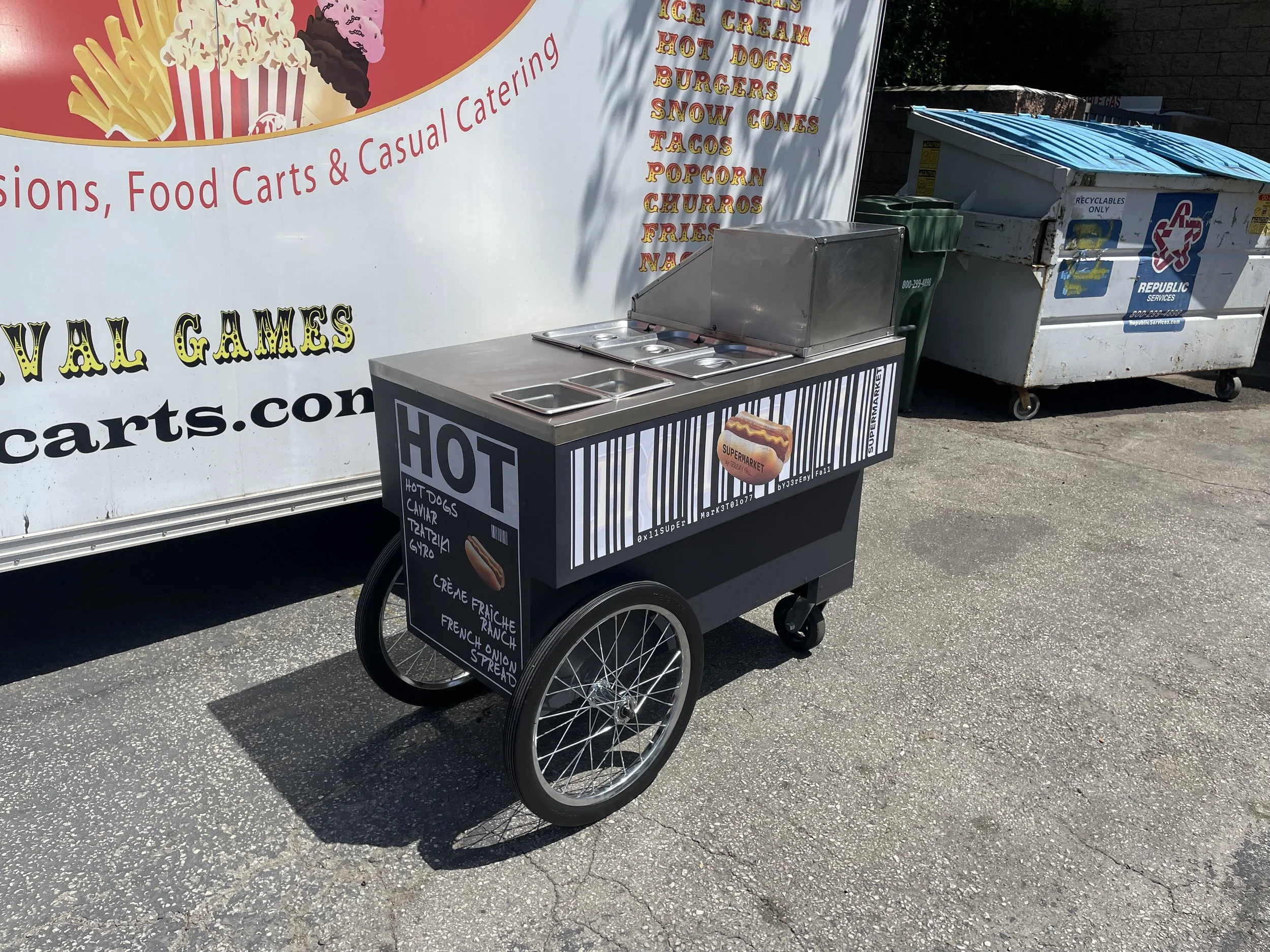 A food cart on a street selling hot dogs, churros, french fries, and other snacks. Behind the cart, there is a large sign advertising food options, and a recycling bin is nearby.