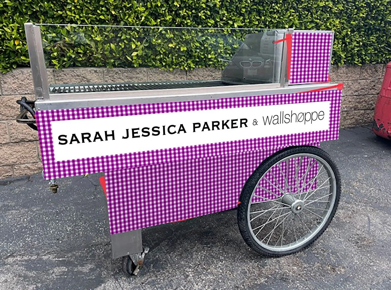 A mobile food cart with a pink and white checkered pattern, displaying a sign that reads 'Sarah Jessica Parker & wallshoppe', parked on an asphalt surface with a brick wall and green bushes in the background.