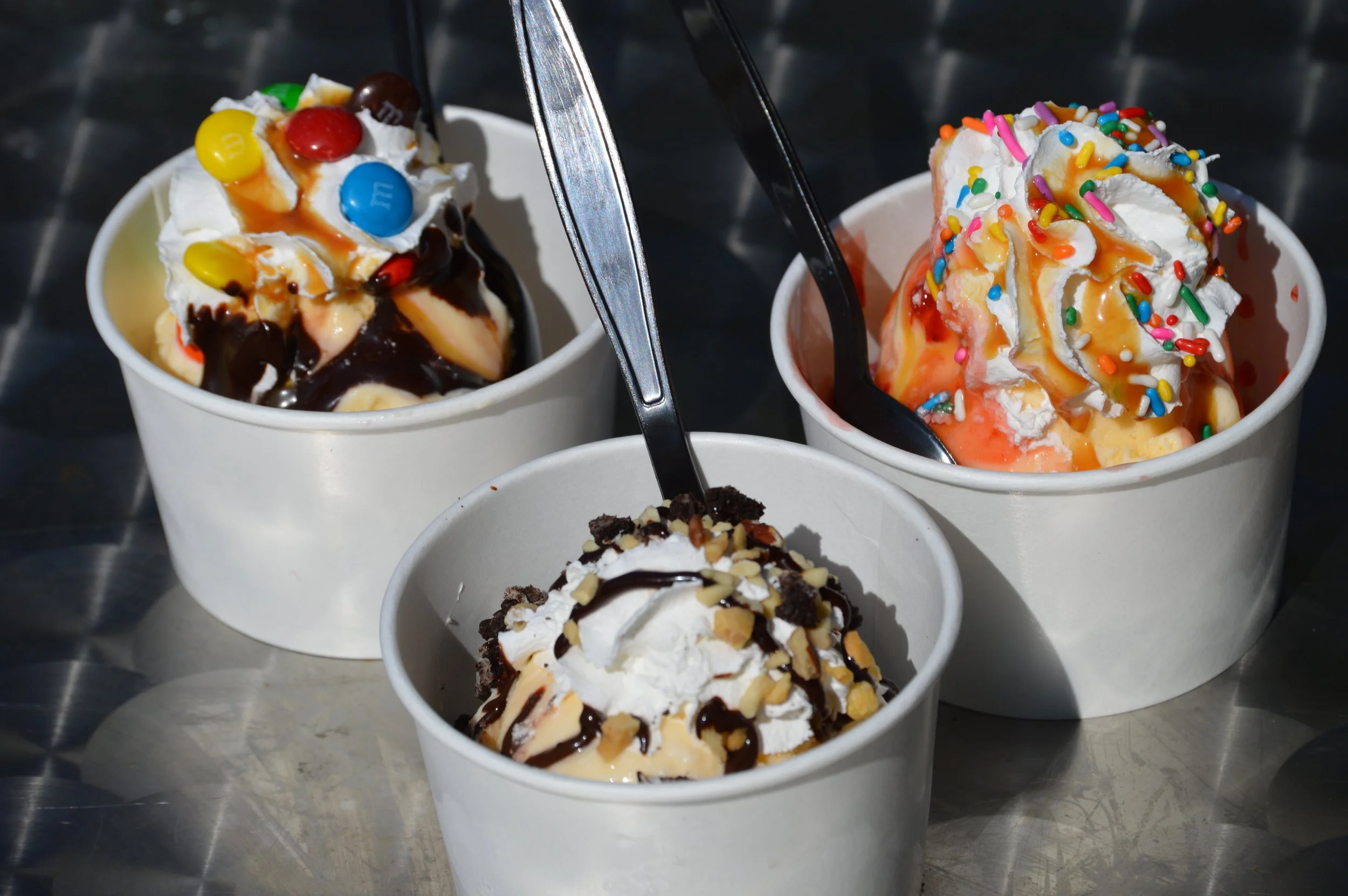Three cups of ice cream with toppings, including chocolate, caramel, nuts, sprinkles, and candies, on a metal surface.