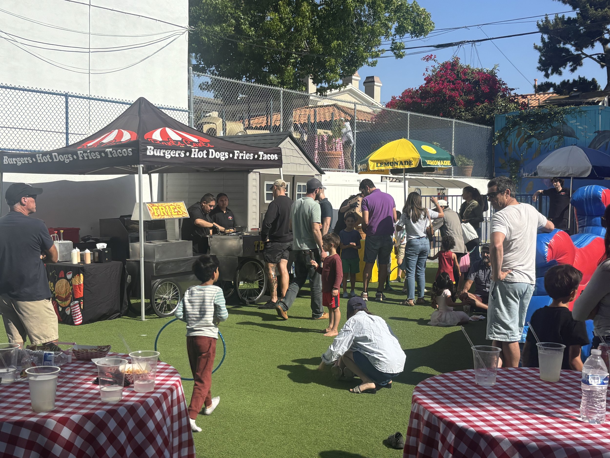 Scene of an outdoor special event with people standing and sitting on artificial grass. Food tables with red and white checkered tablecloths & Food stations serving burgers, hot dogs, fries, tacos, lemonade, and beverages to children playing.