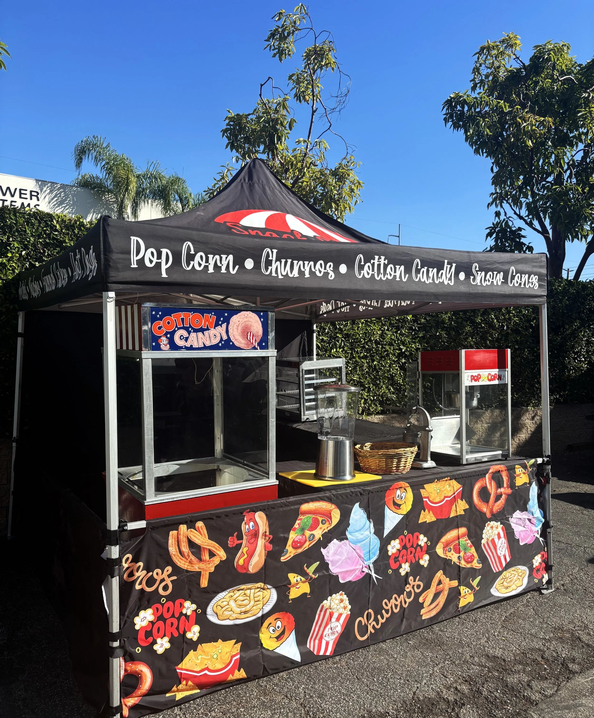 A carnival popcorn stand with a black canopy featuring white text that lists snacks such as pop corn, churros, cotton candy, and snow cones. The front of the stand has colorful illustrations of various carnival treats like pretzels, hot dogs, pizza, 