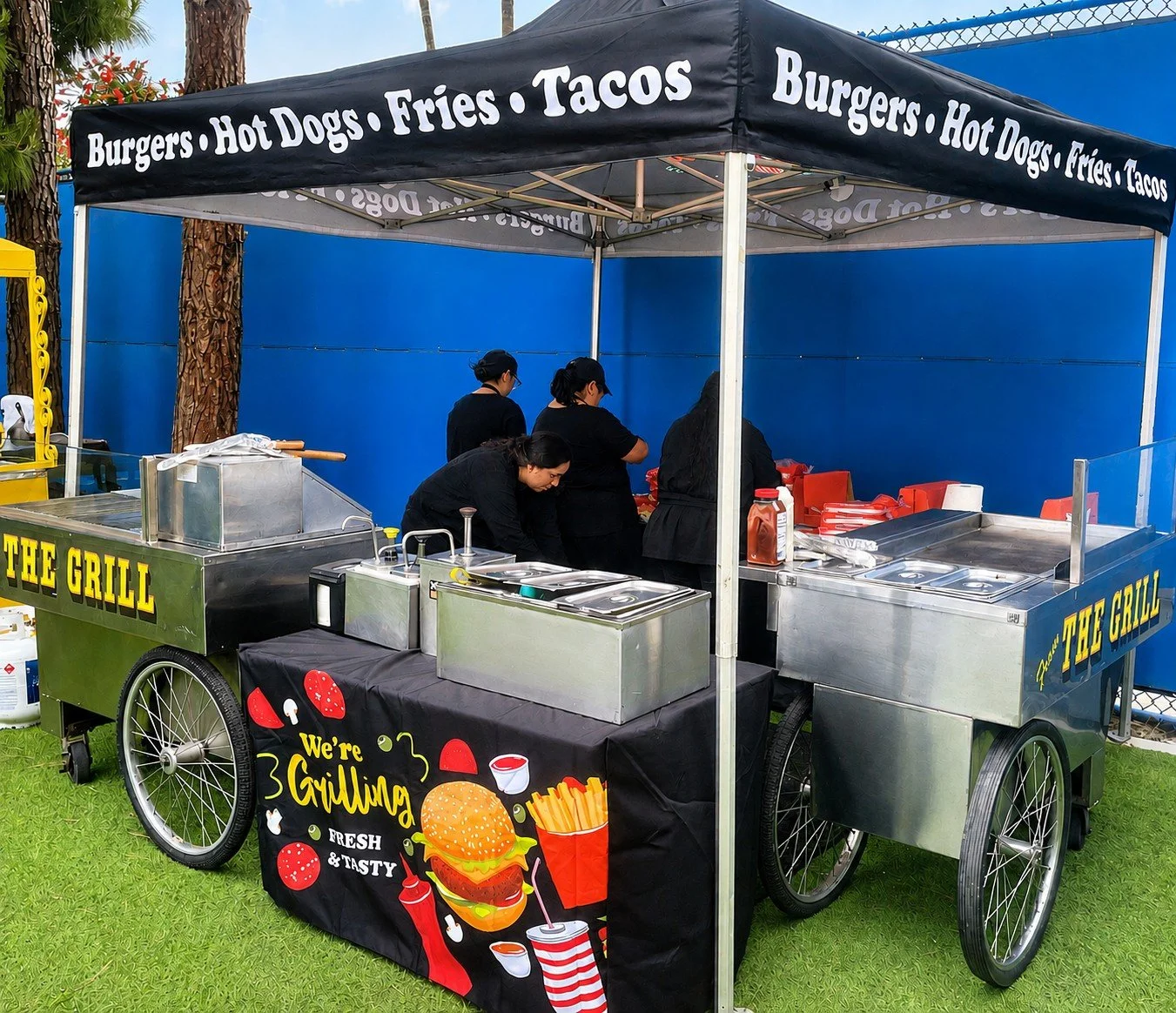 A food stand with a canopy that has text advertising burgers, hot dogs, fries, tacos. The stand has a sign that says 'The Grill' and a sign that says 'We're Grilling, Fresh & Tasty.' There are four people preparing food behind the counter, which has 