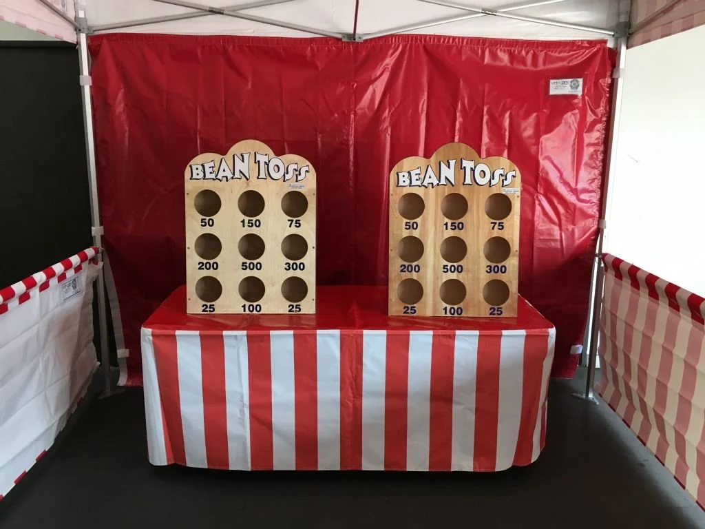 Bean toss game booth with two wooden stands labeled "Bean Toss," each with six circular holes and numbered point values, set up inside a red and white striped tent.