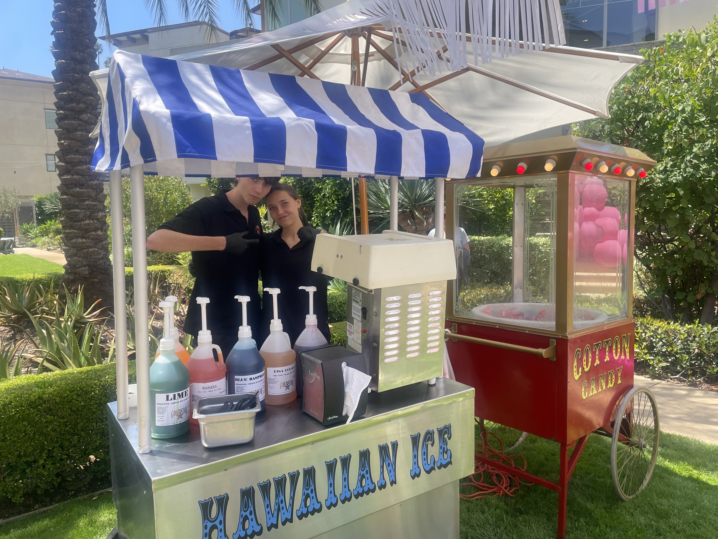 Two young women working at a cotton candy stand outdoors, with containers of colorful flavored syrup in front of them and a red cotton candy machine to the side. There are trees and a building in the background.