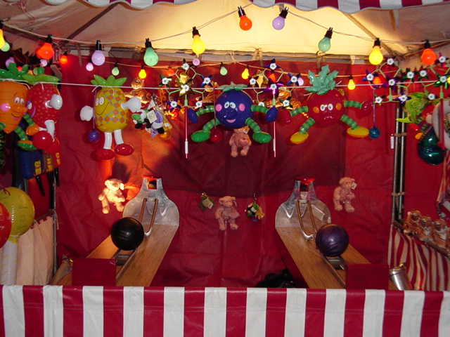 Colorful carnival game booth with stuffed animals, string lights, and plush characters, including a pineapple, blueberry, and strawberry, hanging from the ceiling. The booth has two scoops for knocking down balls.