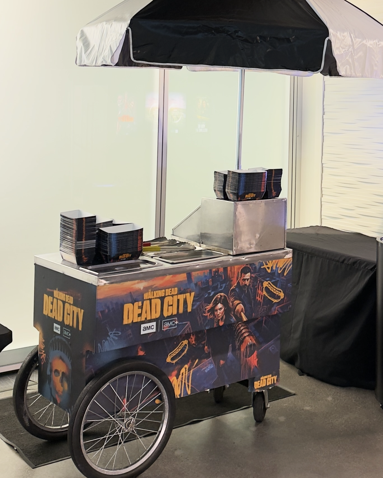A promotional cart for the TV series 'The Walking Dead: Dead City' featuring artwork of characters, stacked black boxes with orange and black advertisements, a metal food container, and a large black umbrella, located indoors next to a white wall with a glass window.