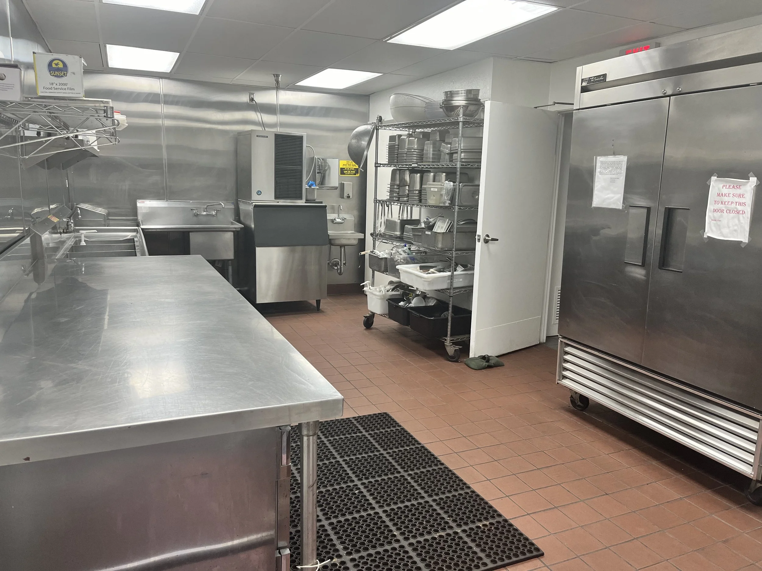 A commercial kitchen with stainless steel equipment, shelving with dishes, a stainless steel sink, a large refrigerator, and a counter. The floor has a textured mat, and a pair of shoes are near the door. Signs are posted on the walls and door.