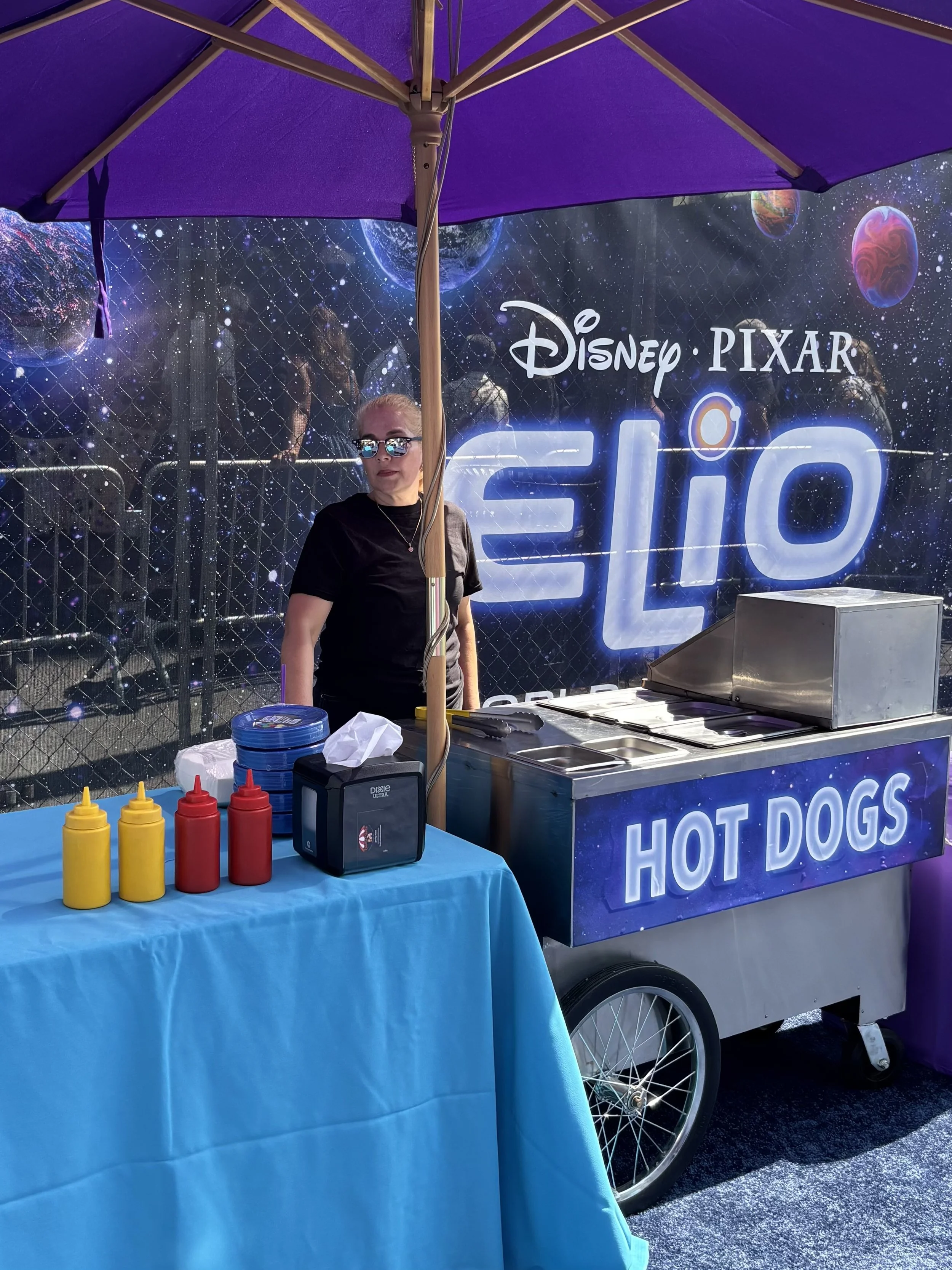 A woman stands behind a hot dog stand at an outdoor event, with purple umbrellas and a backdrop featuring the Disney Pixar logo and space-themed planets and stars. The stand has bottles of mustard and ketchup, and a container labeled 'HOT DOGS.'