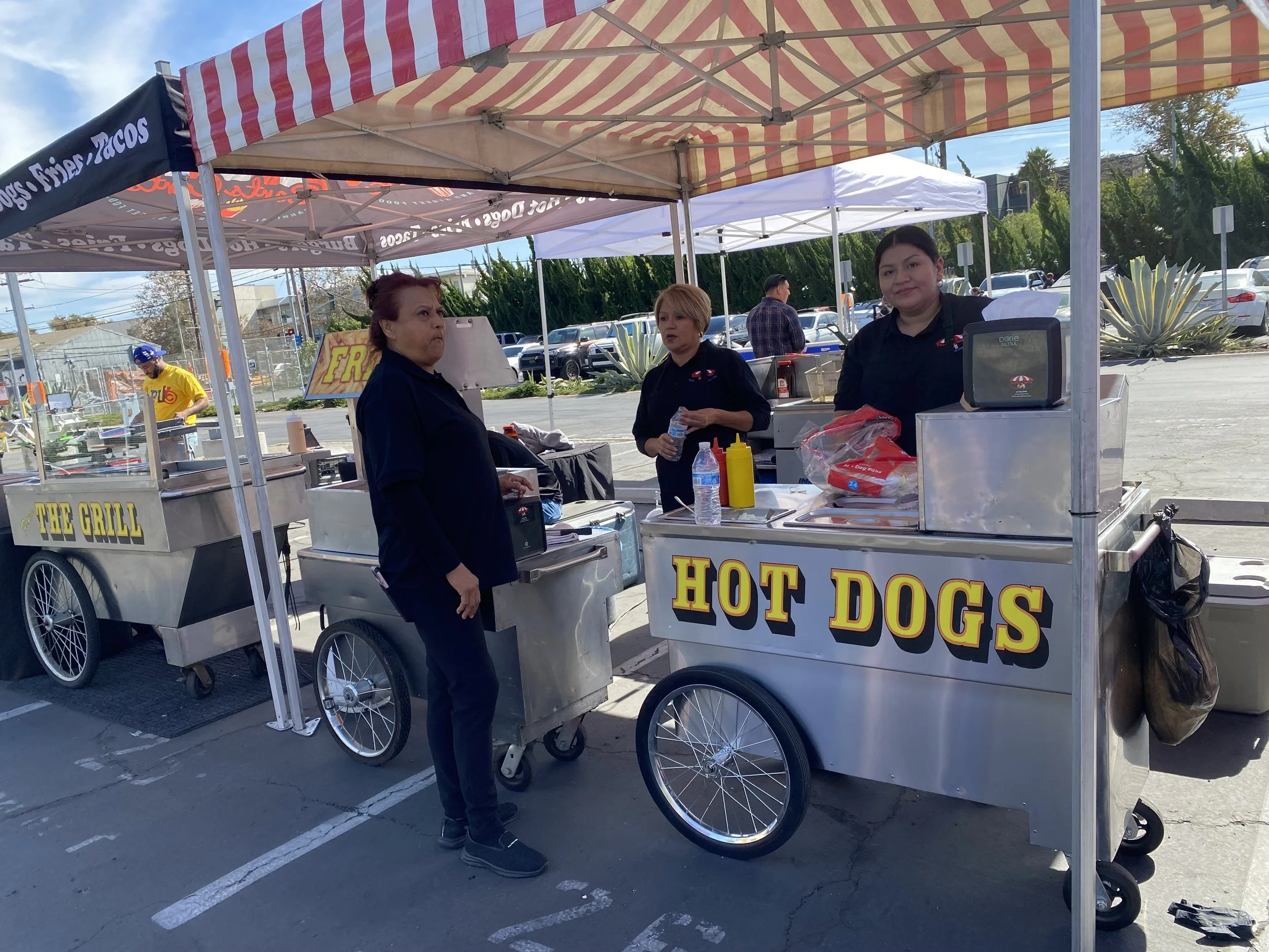 People working at a hot dog stand under striped tents in a parking lot. There is a sign reading 'Hog Dogs' on the stand.