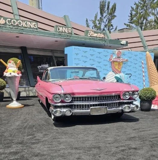 Pink vintage car parked in front of a retro-style restaurant or diner with ice cream decorations and a large ice cream cone prop.
