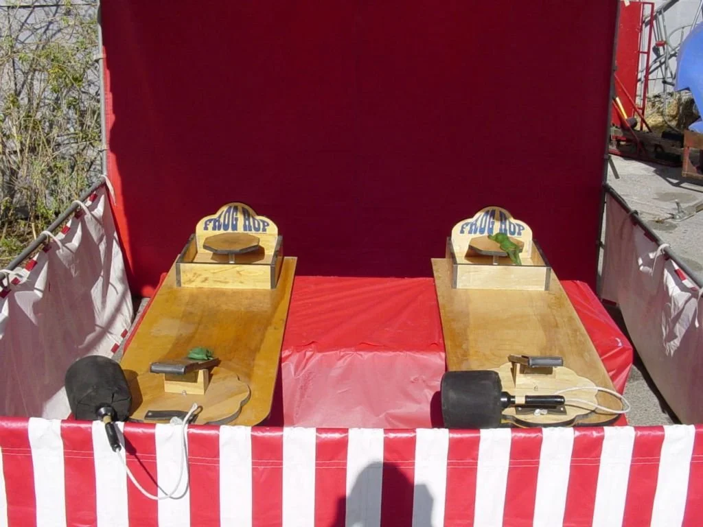 Two wooden skee ball lanes with bumps, each with a ramp and a remote control, set up inside a red and white striped carnival booth with a red backdrop.