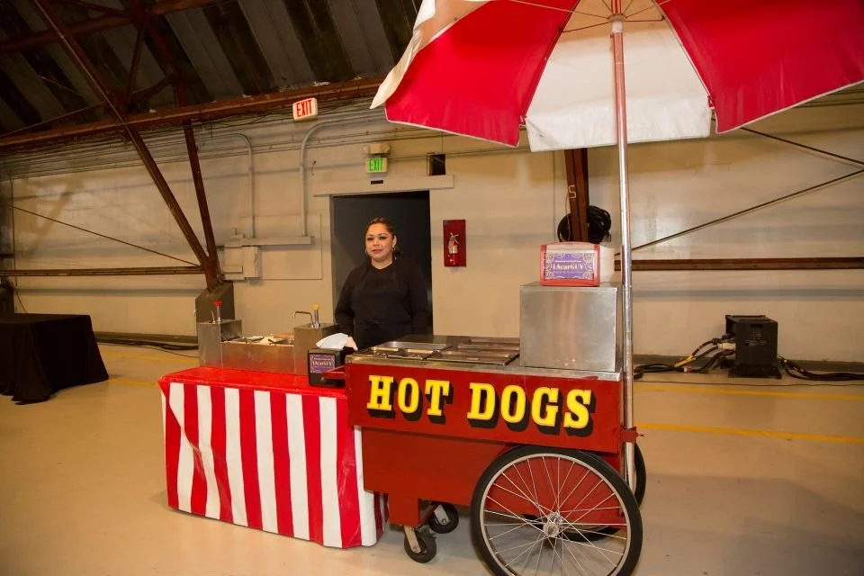 A hot dog stand with a red and white striped tablecloth, located inside a large indoor space. The stand has a red sign with yellow lettering that says 'HOT DOGS' and is equipped with a large red and white umbrella. A woman stands behind the counter.