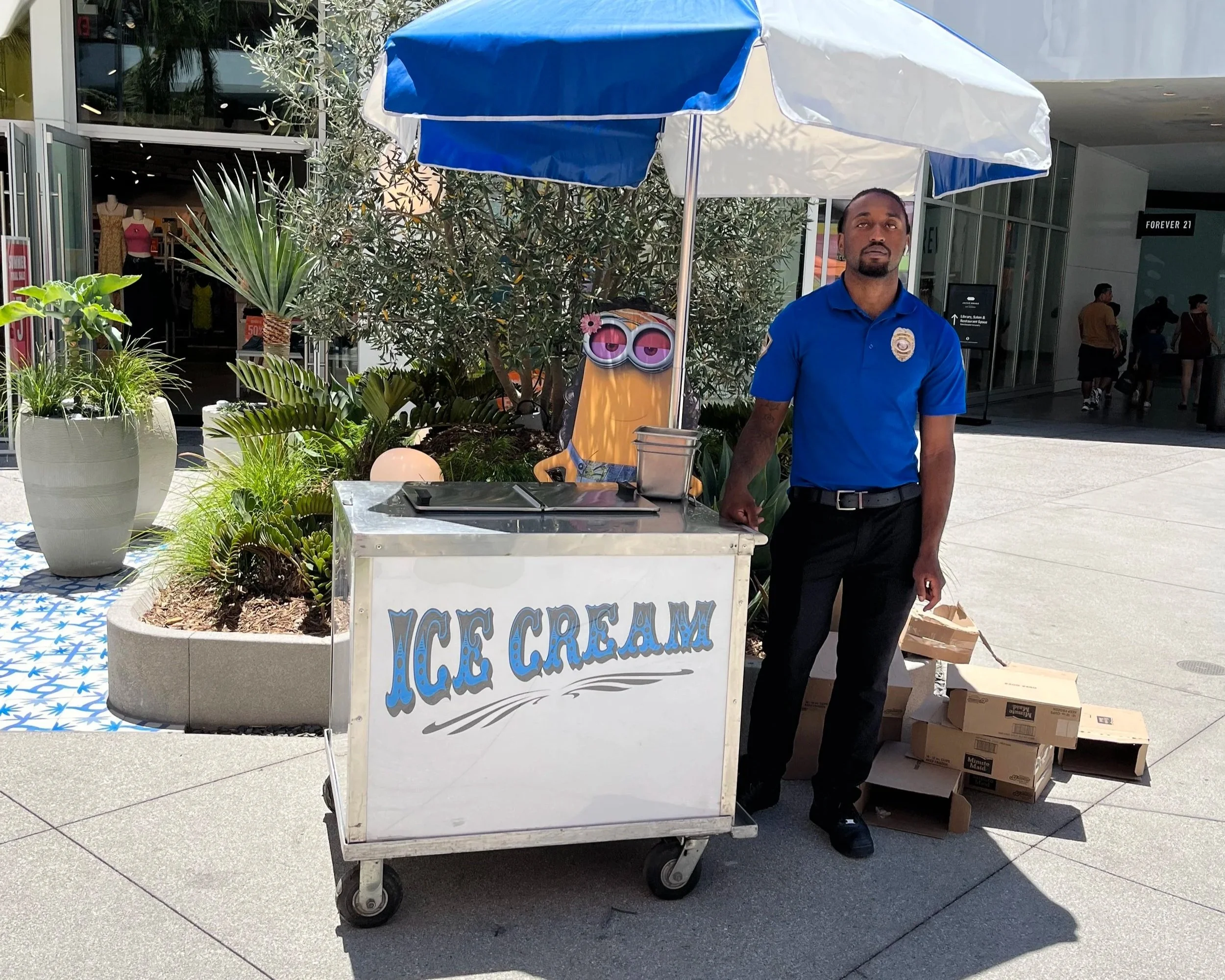 A man wearing a blue security guard uniform standing next to an ice cream cart with a blue and white umbrella. The ice cream cart has a sign that says 'ICE CREAM' and is at an outdoor shopping area with plants and people walking in the background.