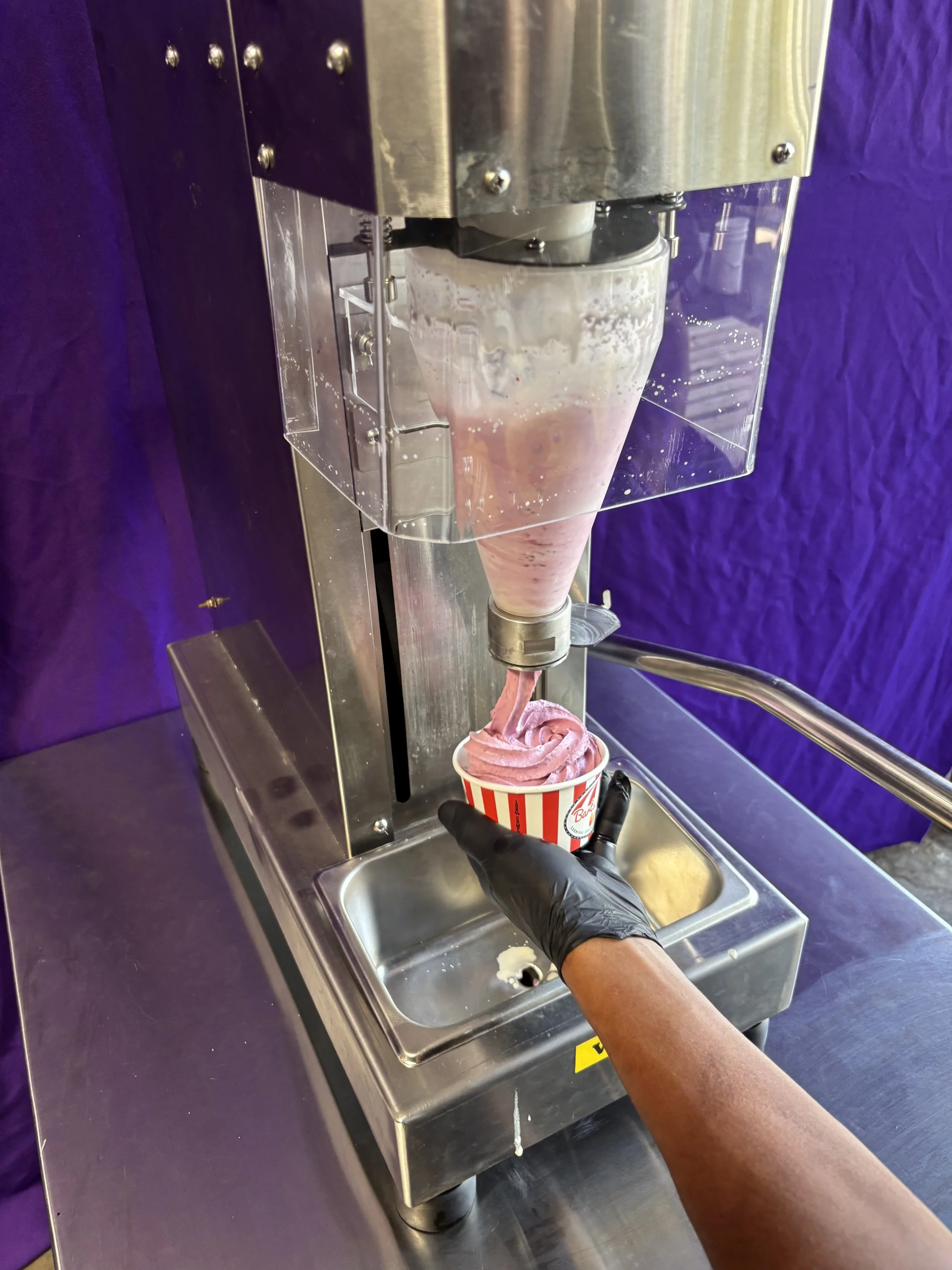 A person wearing a black glove is filling a cup with soft-serve ice cream from an ice cream dispenser over a stainless steel countertop, with a purple wall in the background.