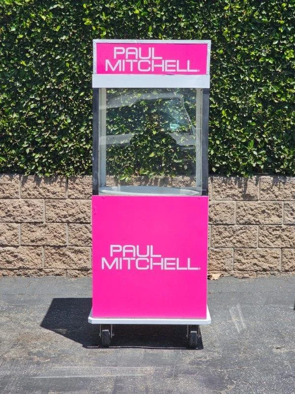 Pink display stand with the name 'Paul Mitchell' on both the top sign and the bottom panel, located outdoors on a sidewalk against a brick and hedge background.