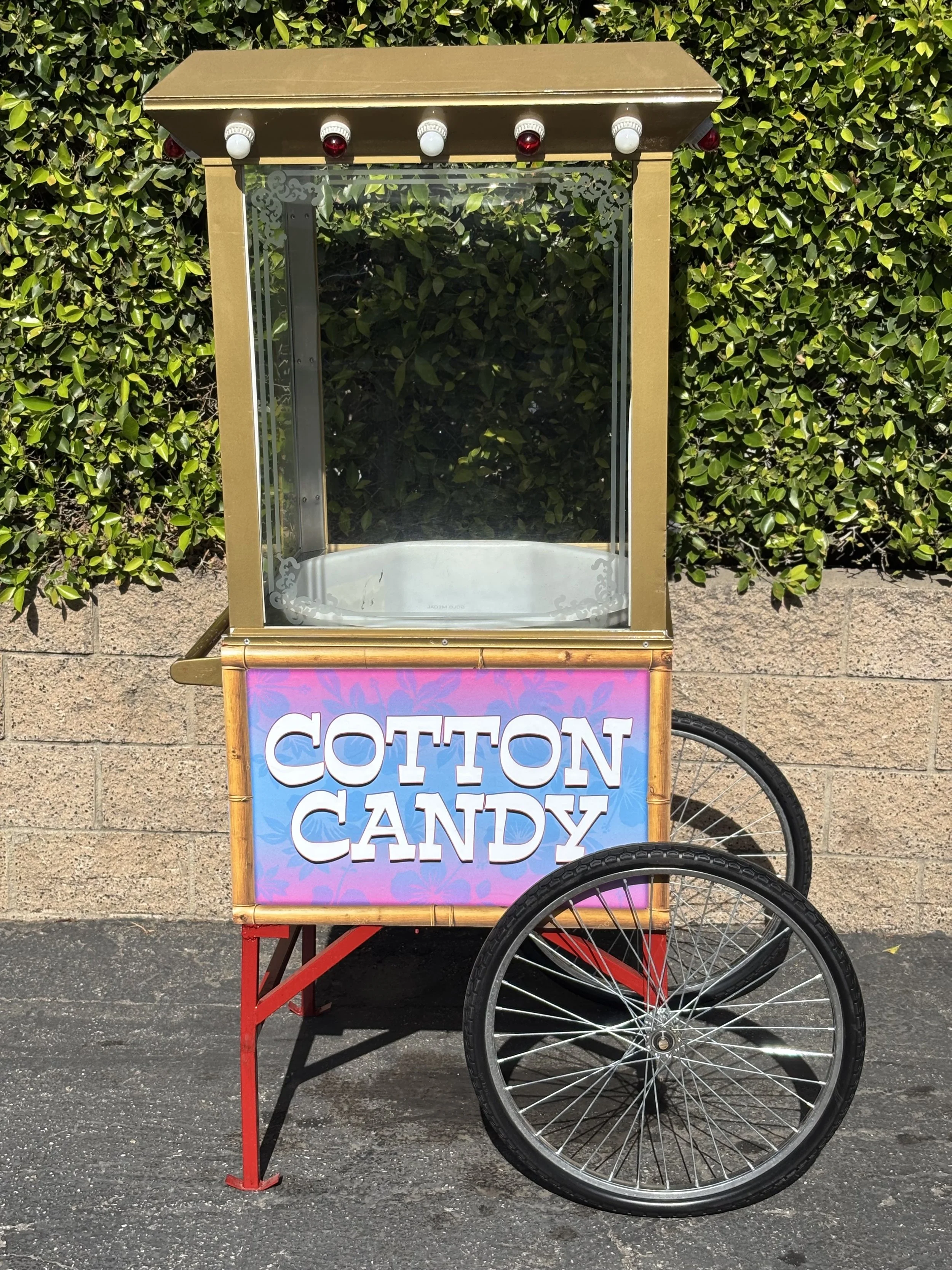 A mobile cotton candy stand in front of a leafy green hedge. The stand has a sign that says 'Cotton Candy' with a pink and blue background, and a clear glass display case on top. The stand is attached to a red bicycle.