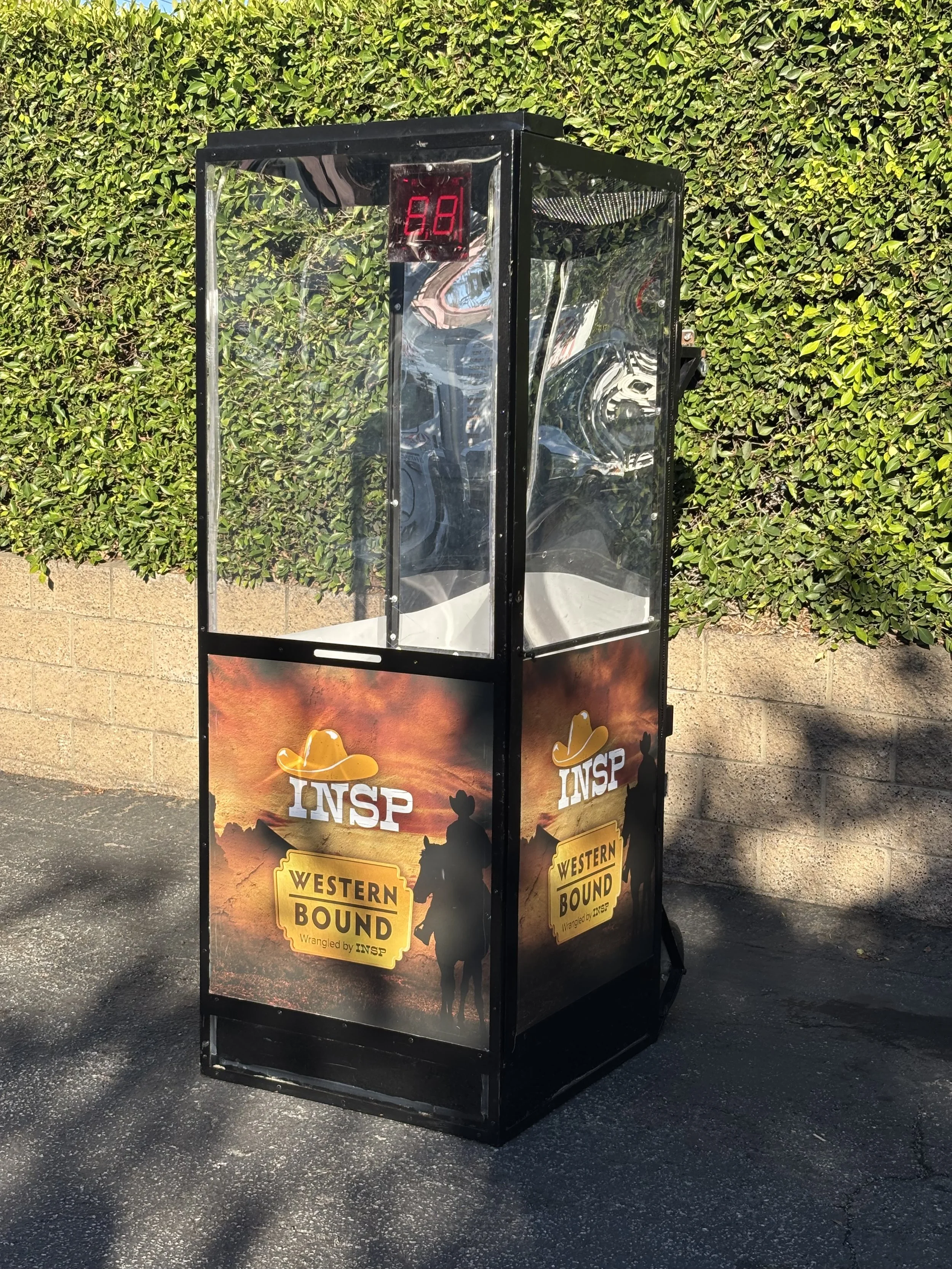 A transparent, rectangular, free-standing display case with a digital timer on the top and a Western-themed display on the lower part, featuring a silhouette of a cowboy and a dog, with the words "INSP" and "Western Bound." The case is positioned outside on asphalt near a brick wall and green hedge.