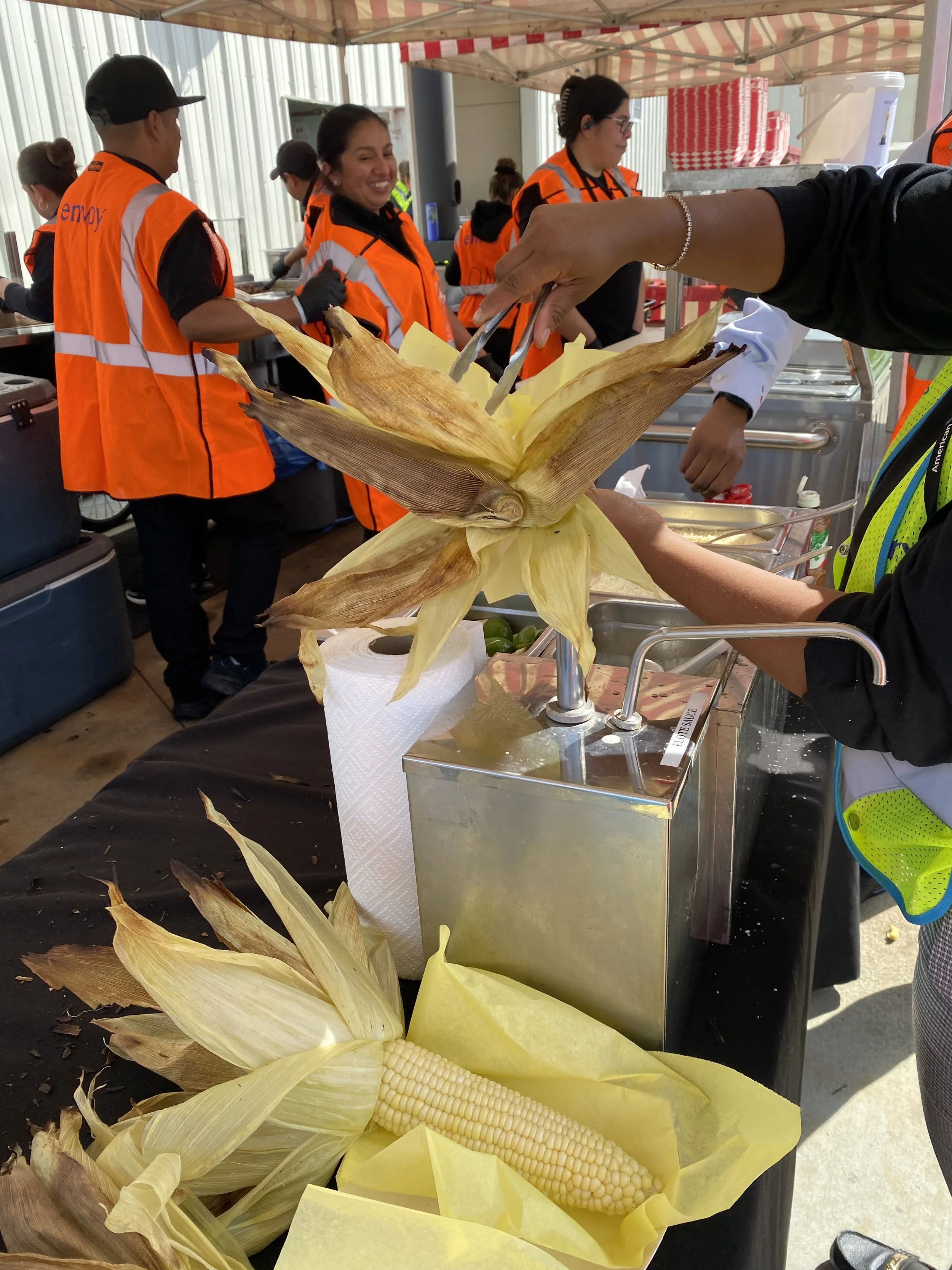 People in orange vests serving food at an outdoor event with corn on the cob in the foreground.