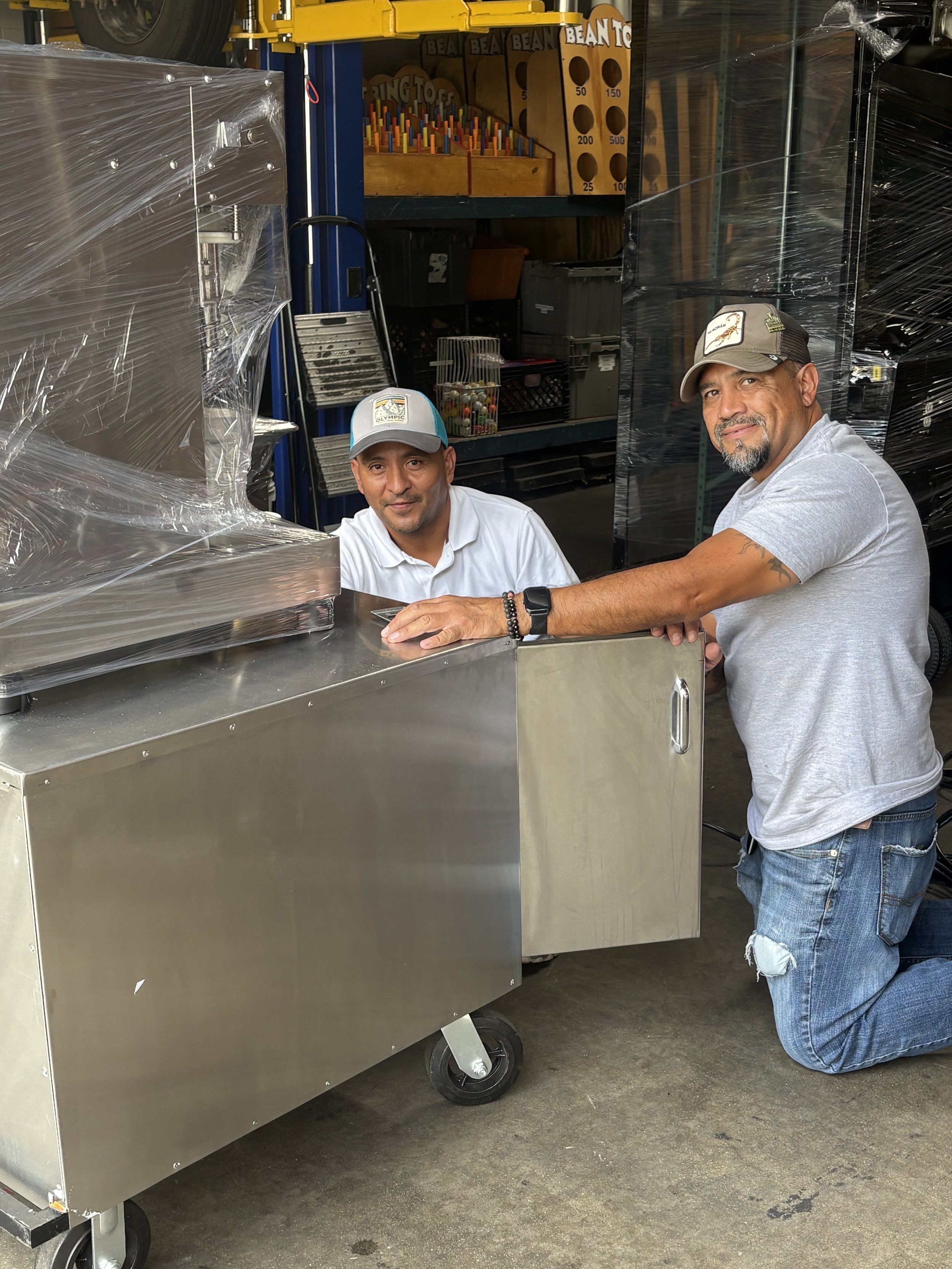 Two men are in a warehouse, one is kneeling and the other is standing behind a shiny metal cart. The man kneeling is wearing a grey t-shirt, ripped jeans, and a baseball cap. The man standing is wearing a white polo shirt and a cap. Shelves with various items including a toy chain, toy peg boards, and boxes can be seen in the background.