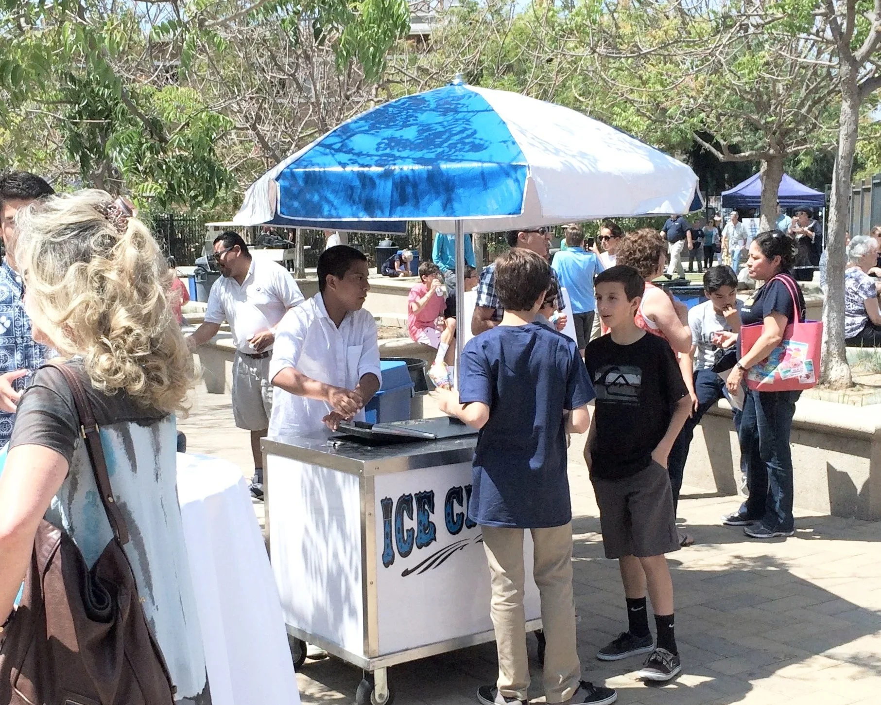 People gathered around a mobile ice cream cart with an umbrella in a park, with trees and other park visitors in the background.