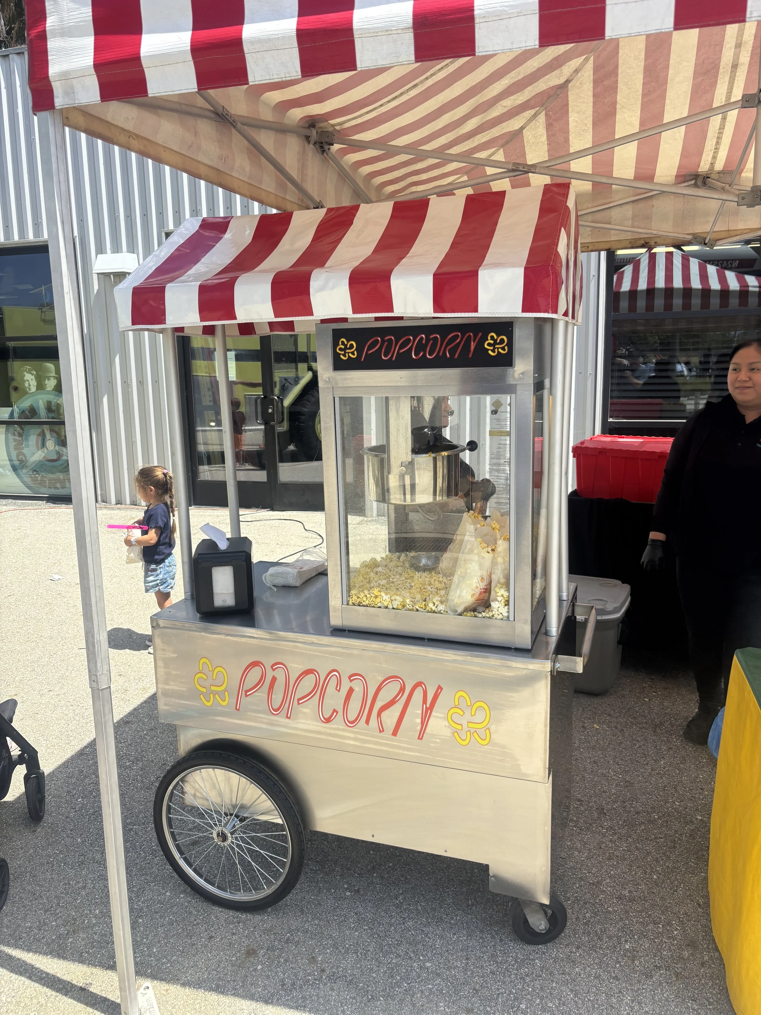 Mobile popcorn stand with a red and white striped canopy and sign, at an outdoor market or fair.