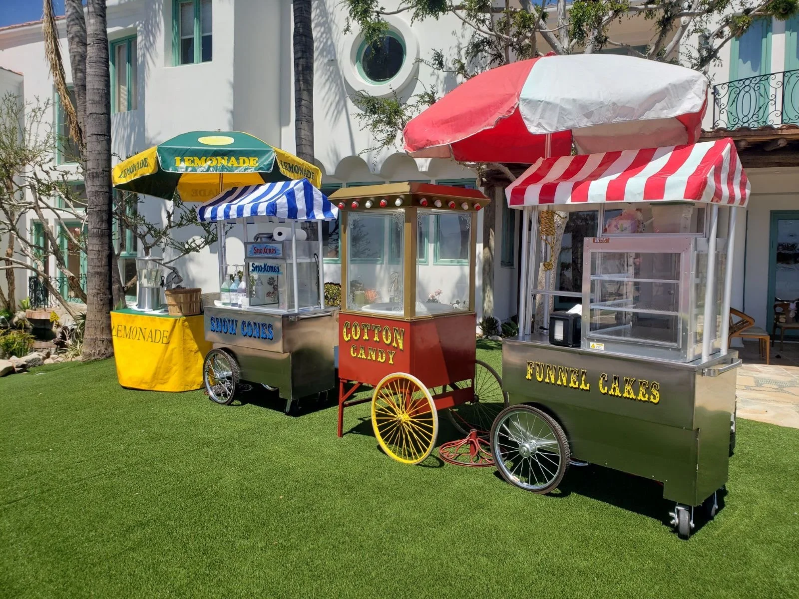 Multiple colorful ice cream and snack stands on a grassy area in front of a white building with palm trees and a balcony. The stands have signage for lemonade, cotton candy, snow cones, funnel cakes, and other treats, with umbrellas providing shade.