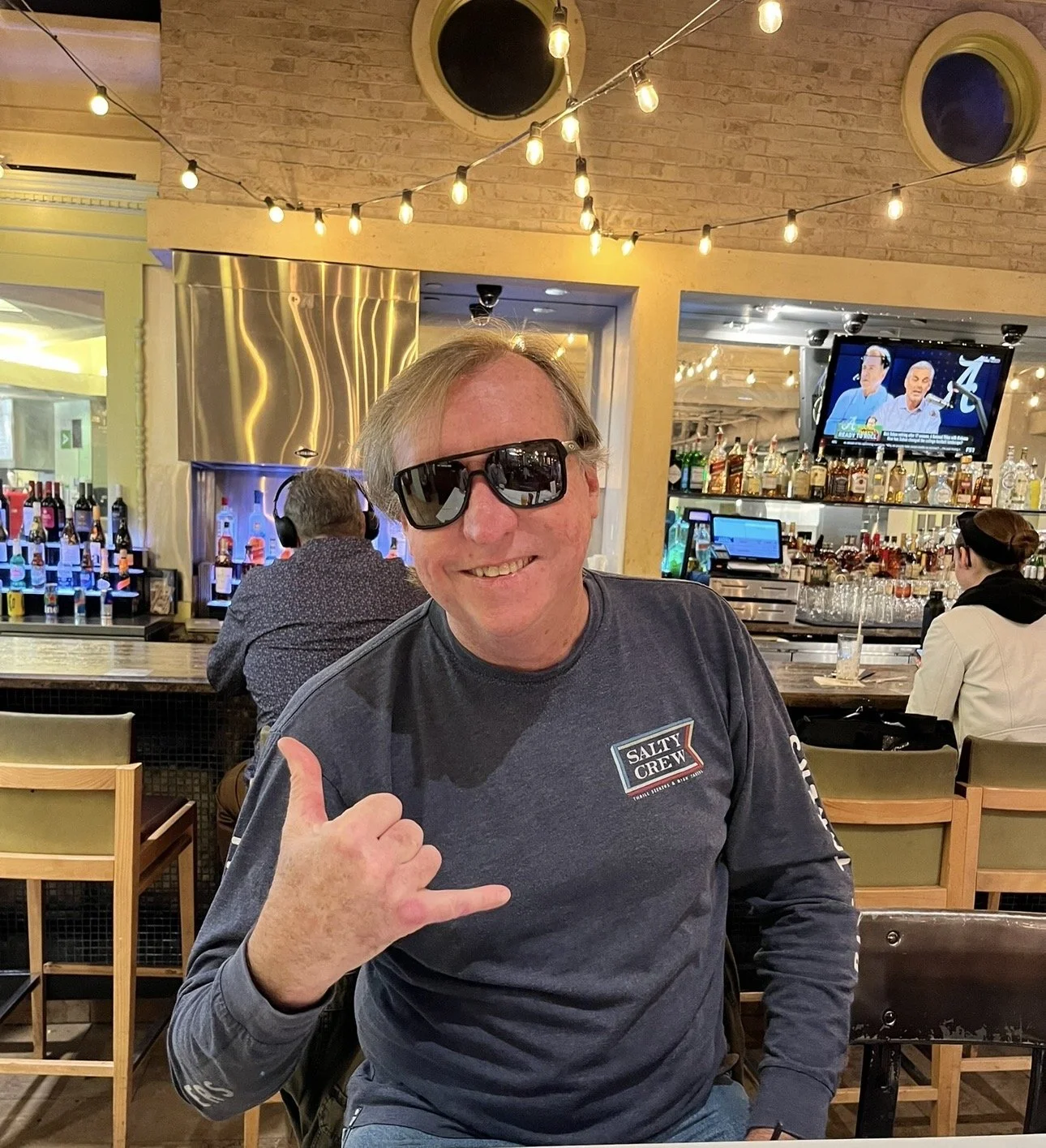 A smiling man wearing sunglasses and a Salty Crew t-shirt making a shaka hand gesture inside a bar or restaurant. There are patrons at the bar and a television in the background.