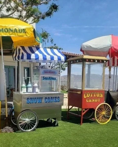 Two vintage-style popcorn and cotton candy carts with colorful umbrellas on a grassy area, outdoors under a clear blue sky.