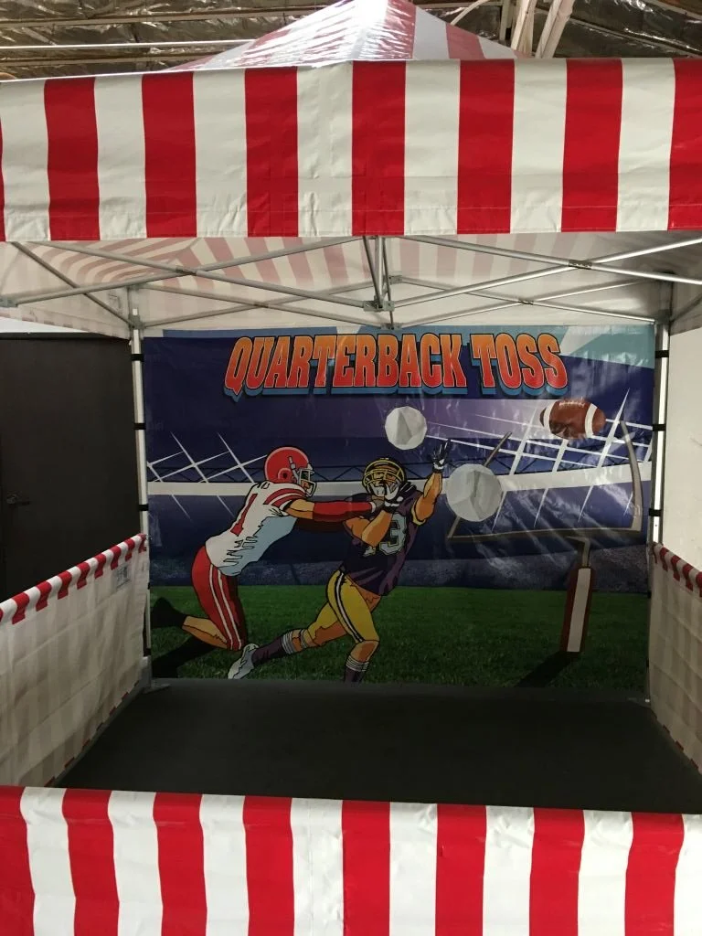 A football game-themed carnival game booth with a banner reading 'Quarterback Toss' showing an illustration of a football player throwing the ball at a target with dummies.