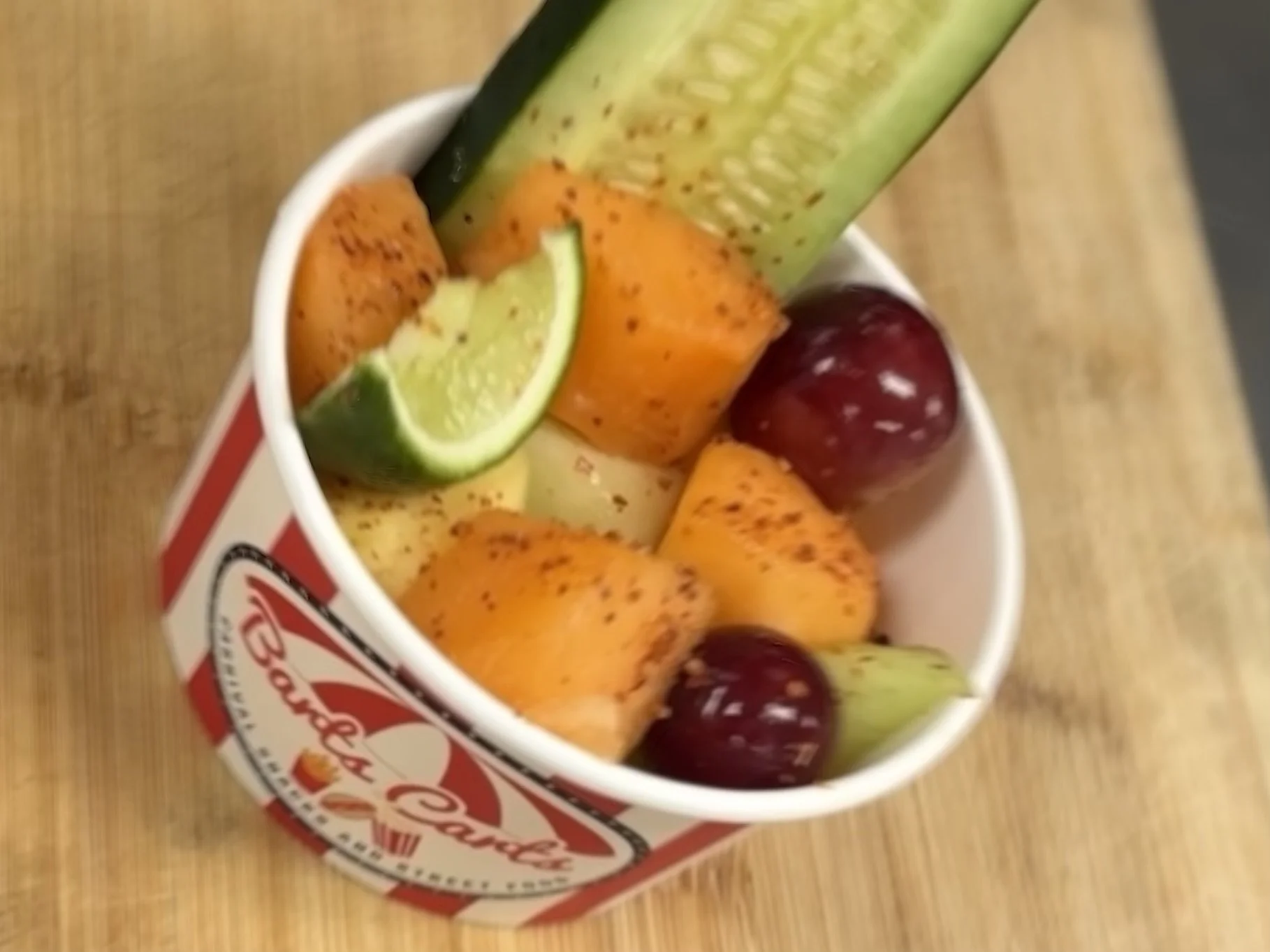 A cup of fruit topped with a lime wedge, pineapple, cherry, and a cucumber slice, sprinkled with black pepper.